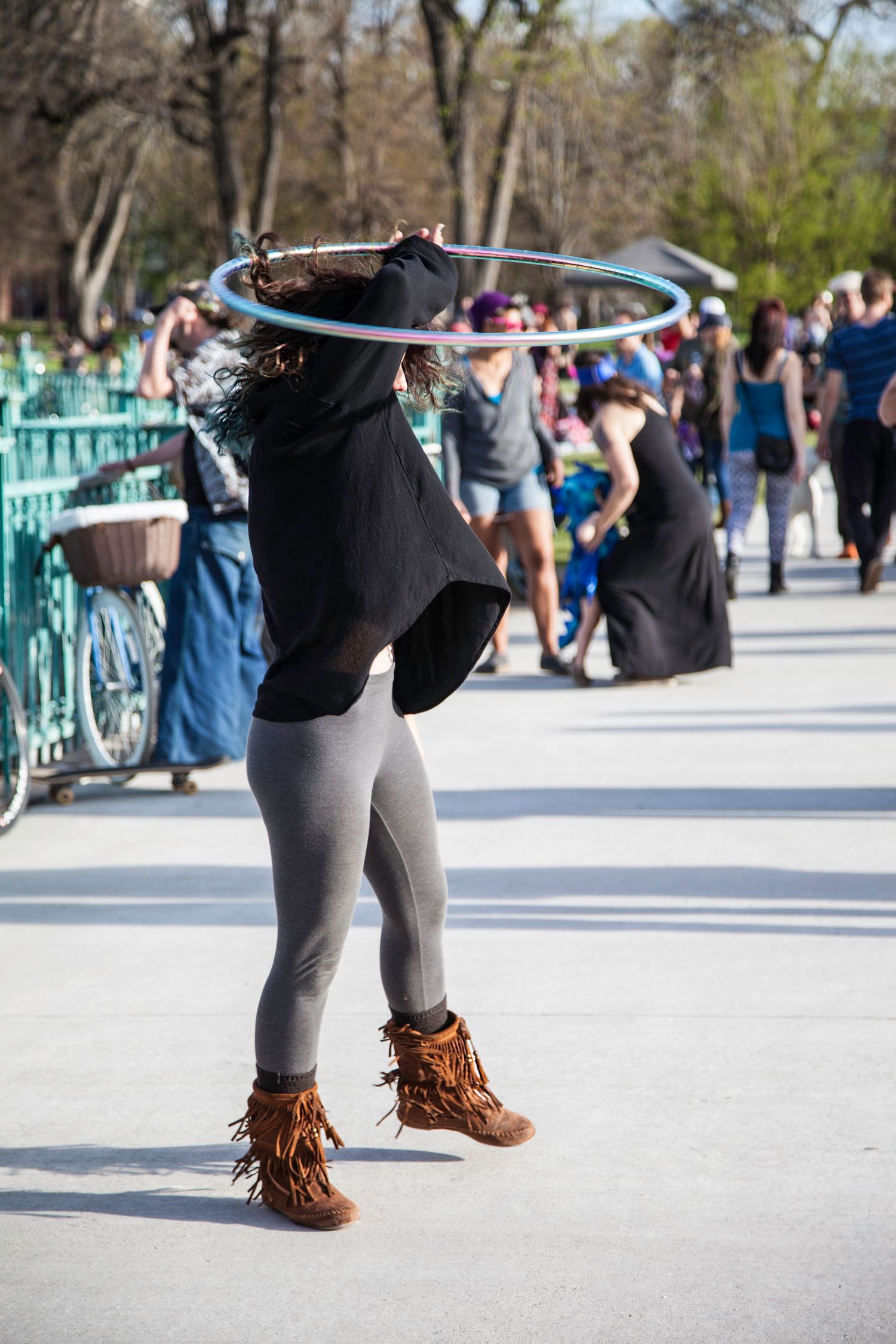 A woman hula hooping with a blue hoop at an outdoor public event in fringed boots.