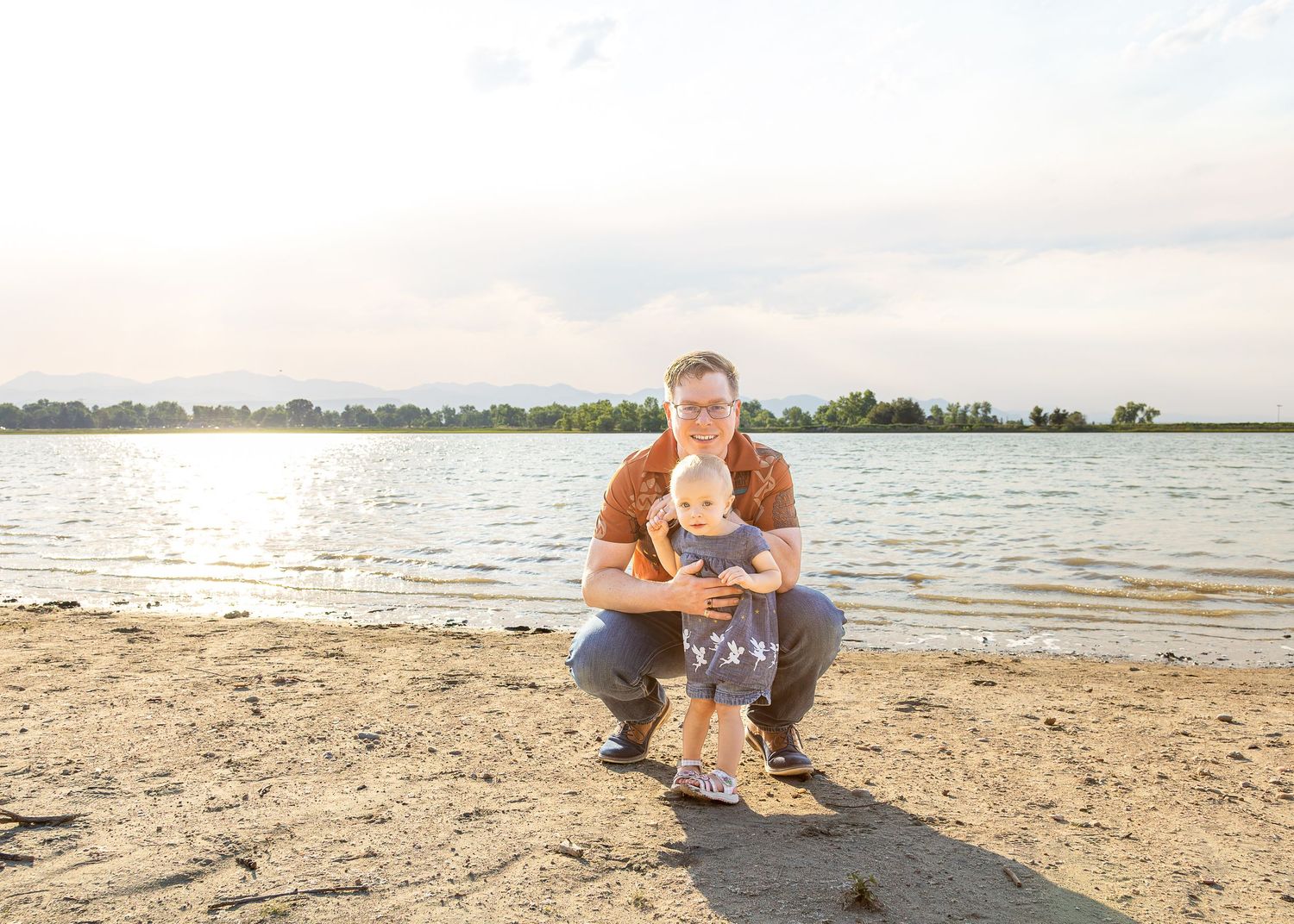 A father crouches on a sandy beach holding his baby with a serene lake and golden sunlight in the background.