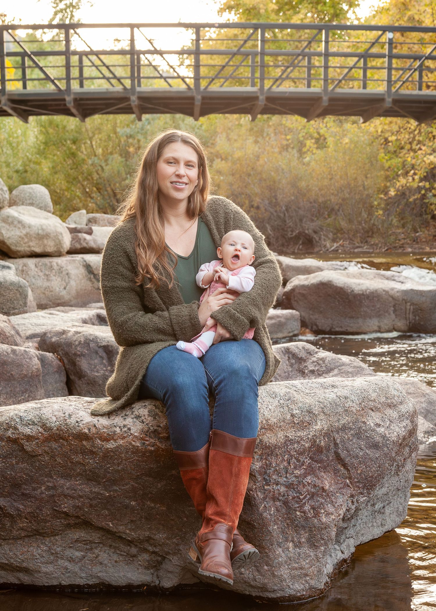 Woman in green cardigan and red boots holds yawning baby on rocks by a creek with a wooden bridge behind.