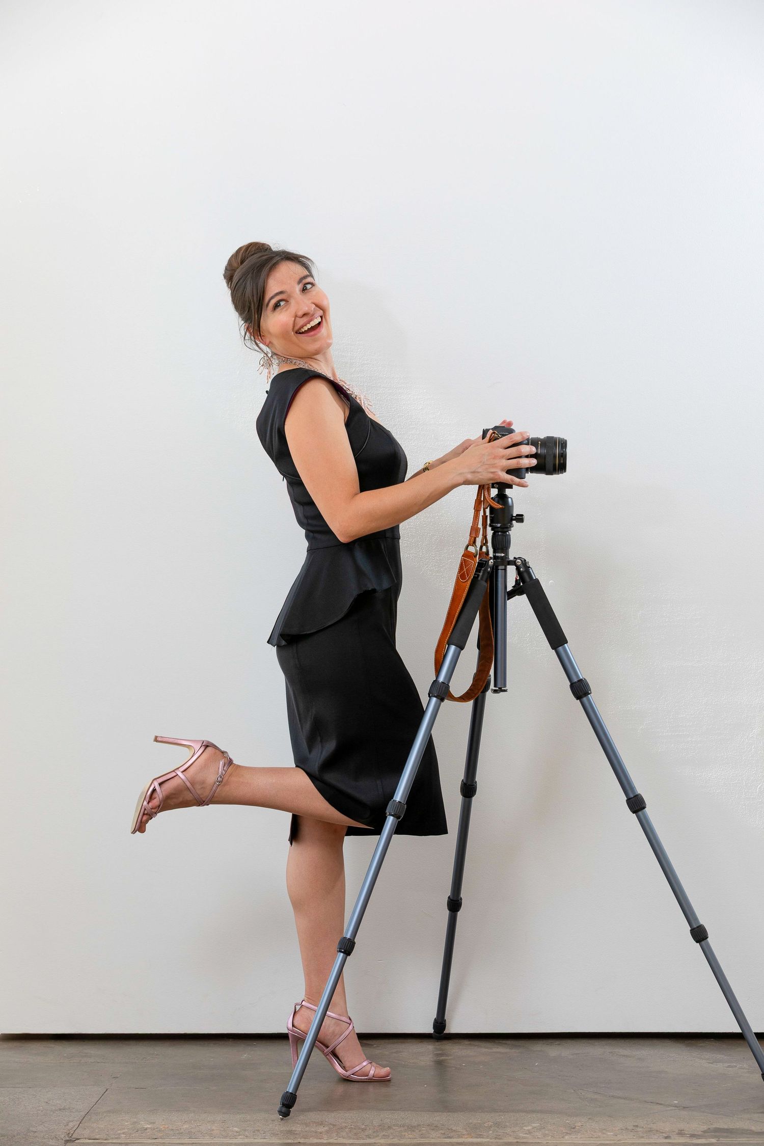 A smiling woman in a black dress and heels poses playfully with a camera on a tripod against a white wall.