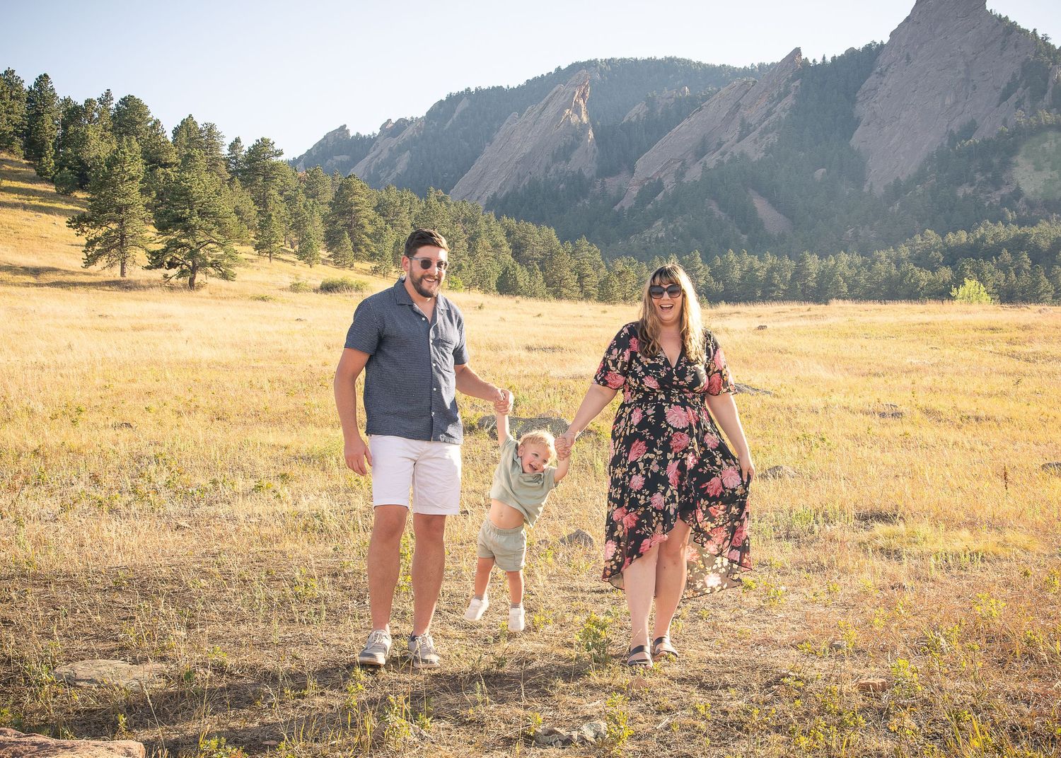 Family of three walking in golden meadow with iconic Flatiron rock formations and pine trees in Boulder, Colorado.