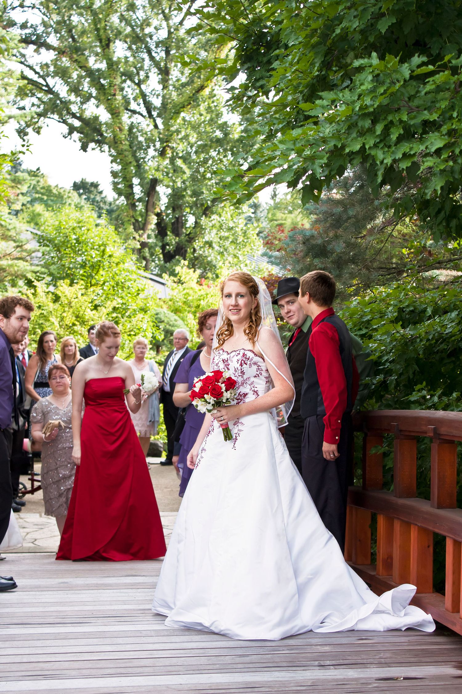 Bride in white gown holds red bouquet on wooden bridge, with wedding party and guests in lush garden backdrop.