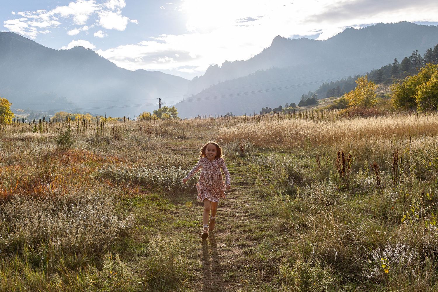 Young girl running through a sunlit mountain meadow with dramatic peaks in the background.