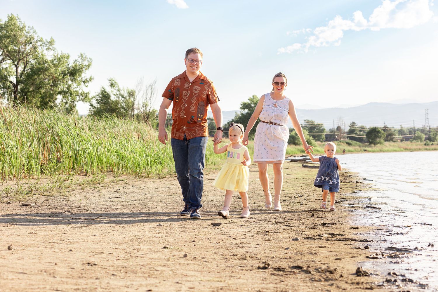 Family of four walking hand-in-hand along a sunny sandy beach shoreline with green reeds and mountains in background.