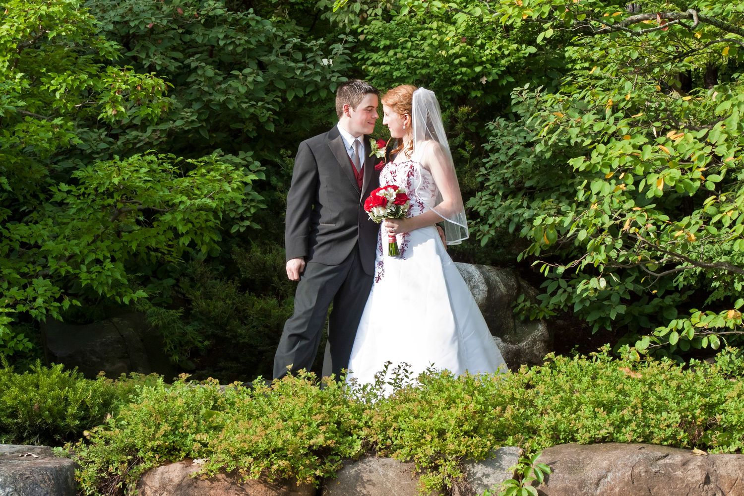 Bride and groom share a romantic moment surrounded by lush green trees, bride holding a red bouquet in a white gown.