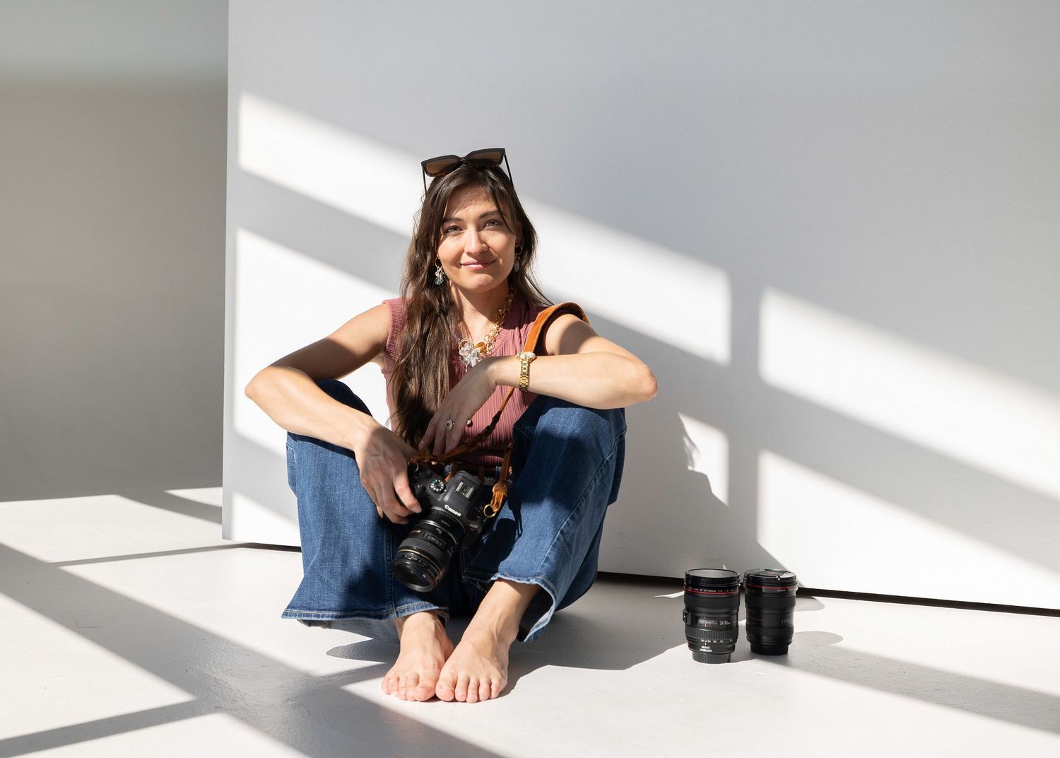 Woman photographer sitting on floor holding DSLR camera, smiling, wearing jeans and tank top with sunglasses on head.