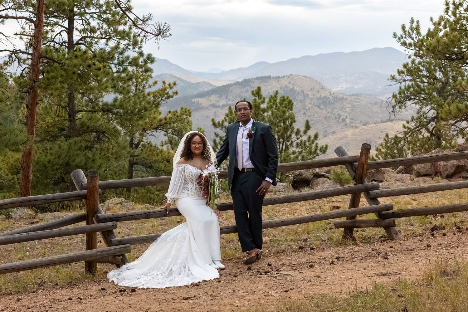 Bride and groom pose by rustic fence with mountain views and pine trees in Colorado wedding photo.