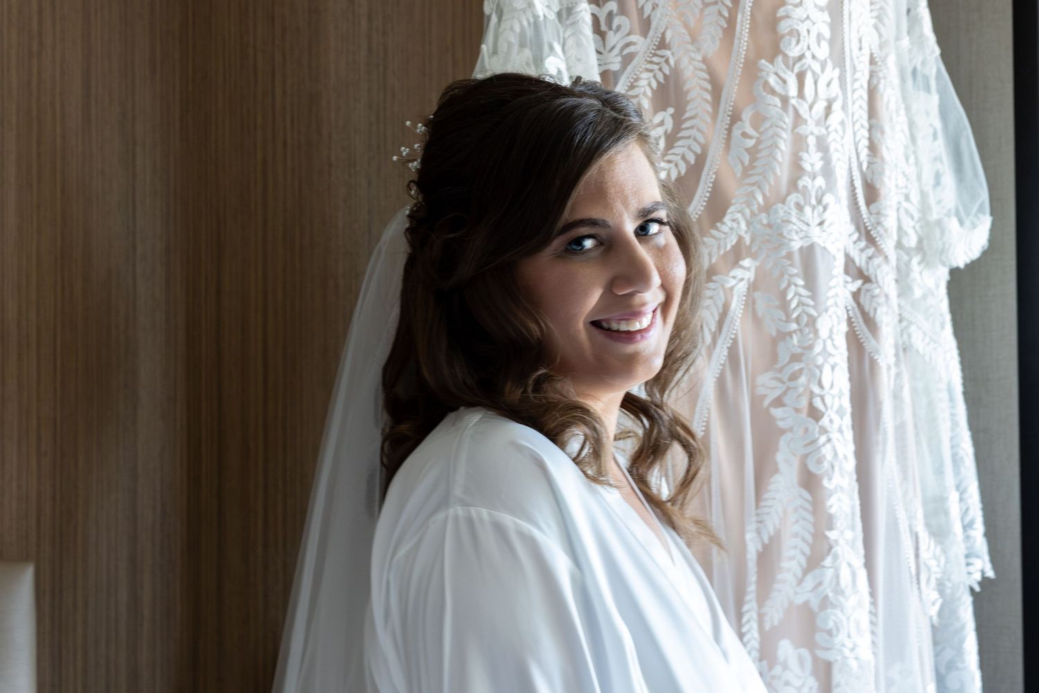 Smiling bride wearing a white robe and veil with lace wedding dress hanging in background.