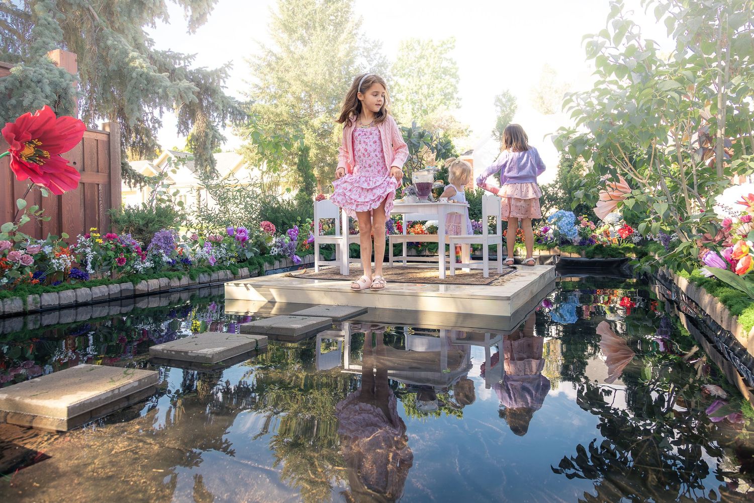 Two young girls in pink dresses stand on a floating platform beside a table surrounded by colorful flowers reflected in a calm garden pond.