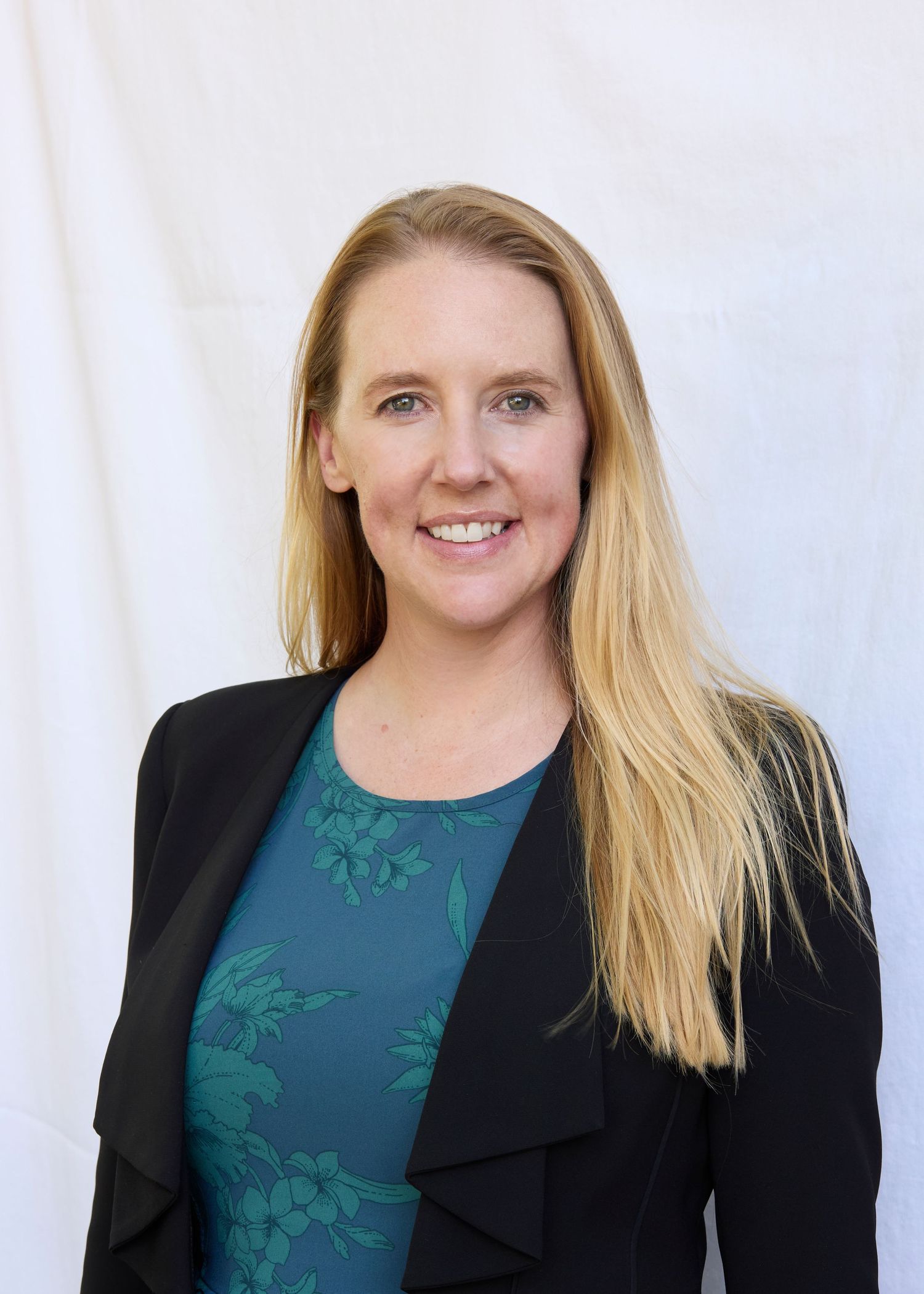 Blonde woman smiling in black blazer and teal floral top against white background.