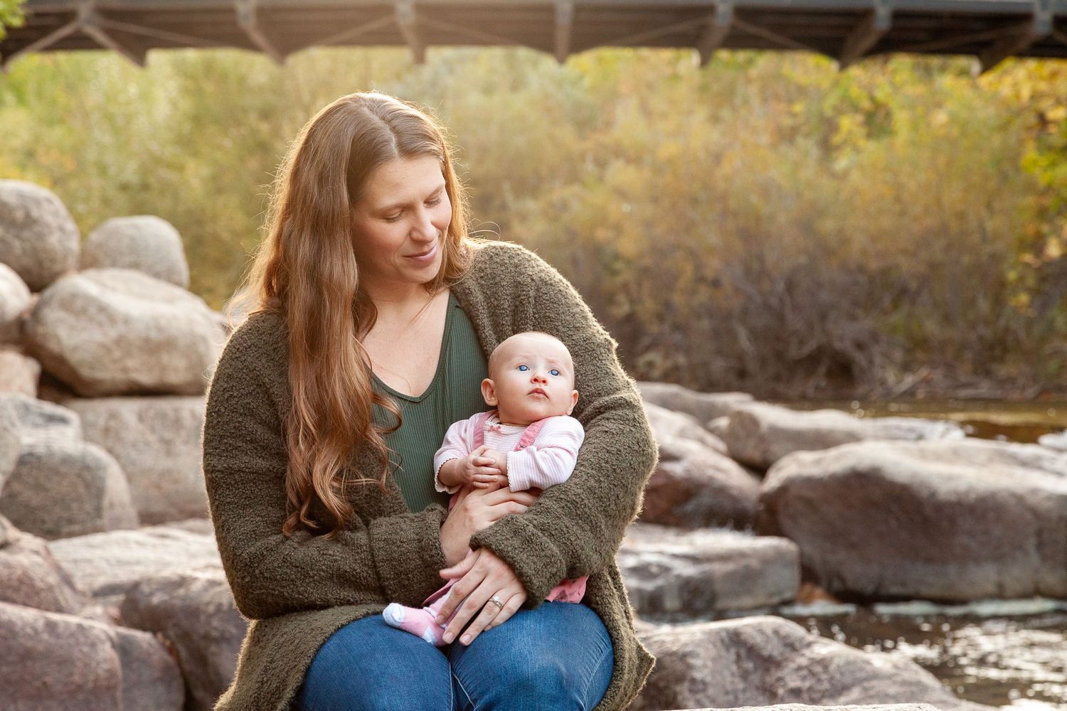 Young mother with long brown hair holds newborn baby outdoors near rocky riverbank with autumn trees.