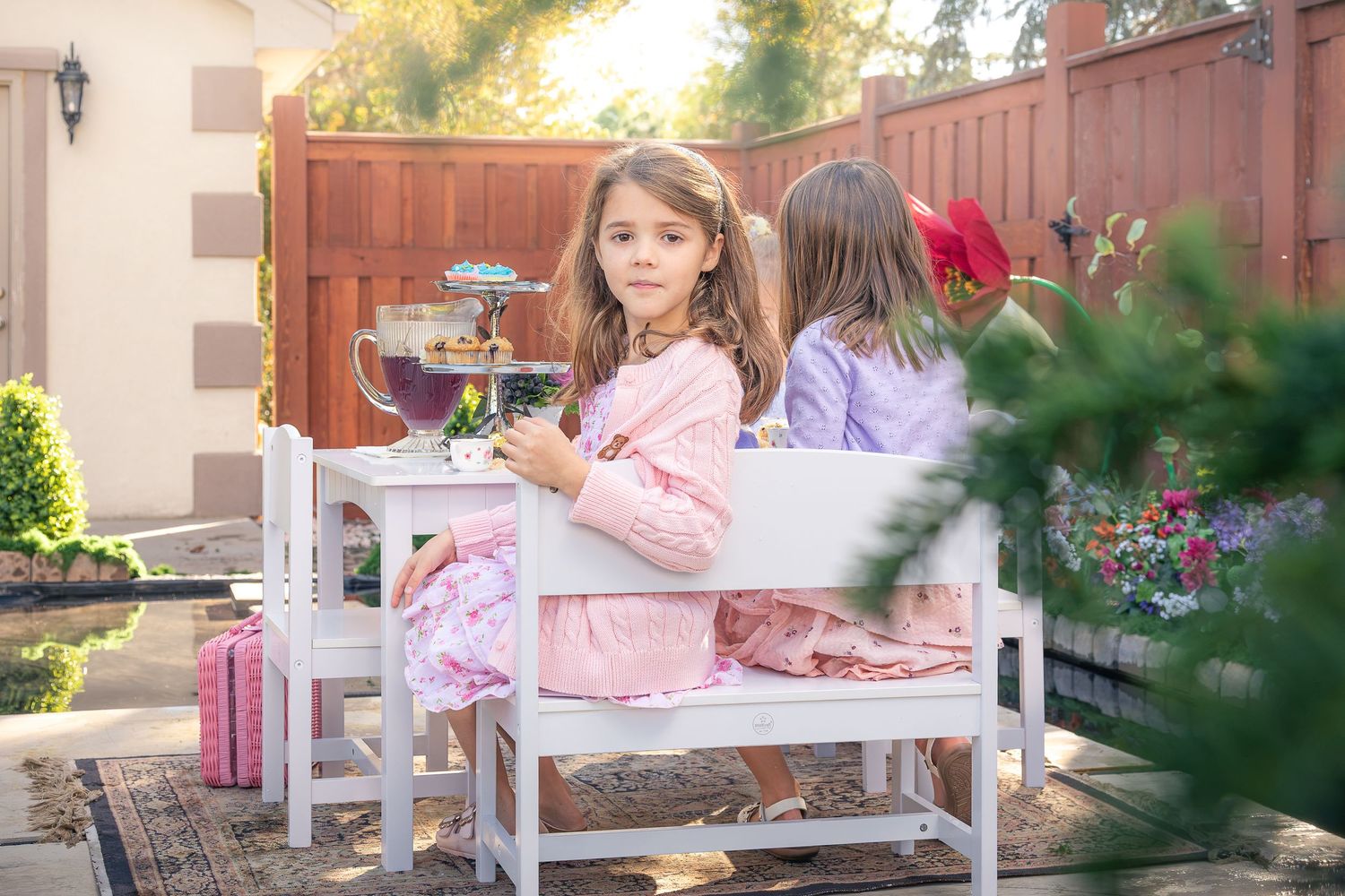 Two young girls in pink and lavender dresses sit at a white child-sized table set for a tea party in a sunny backyard garden.