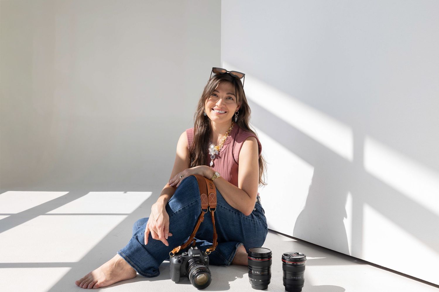 Woman photographer sitting on white studio floor smiling with DSLR camera and lenses beside her.