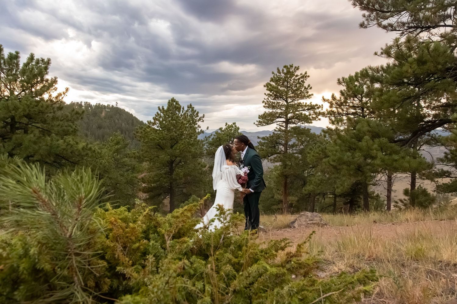 Bride and groom share a romantic moment surrounded by pine trees with dramatic cloudy skies at a mountain wedding.