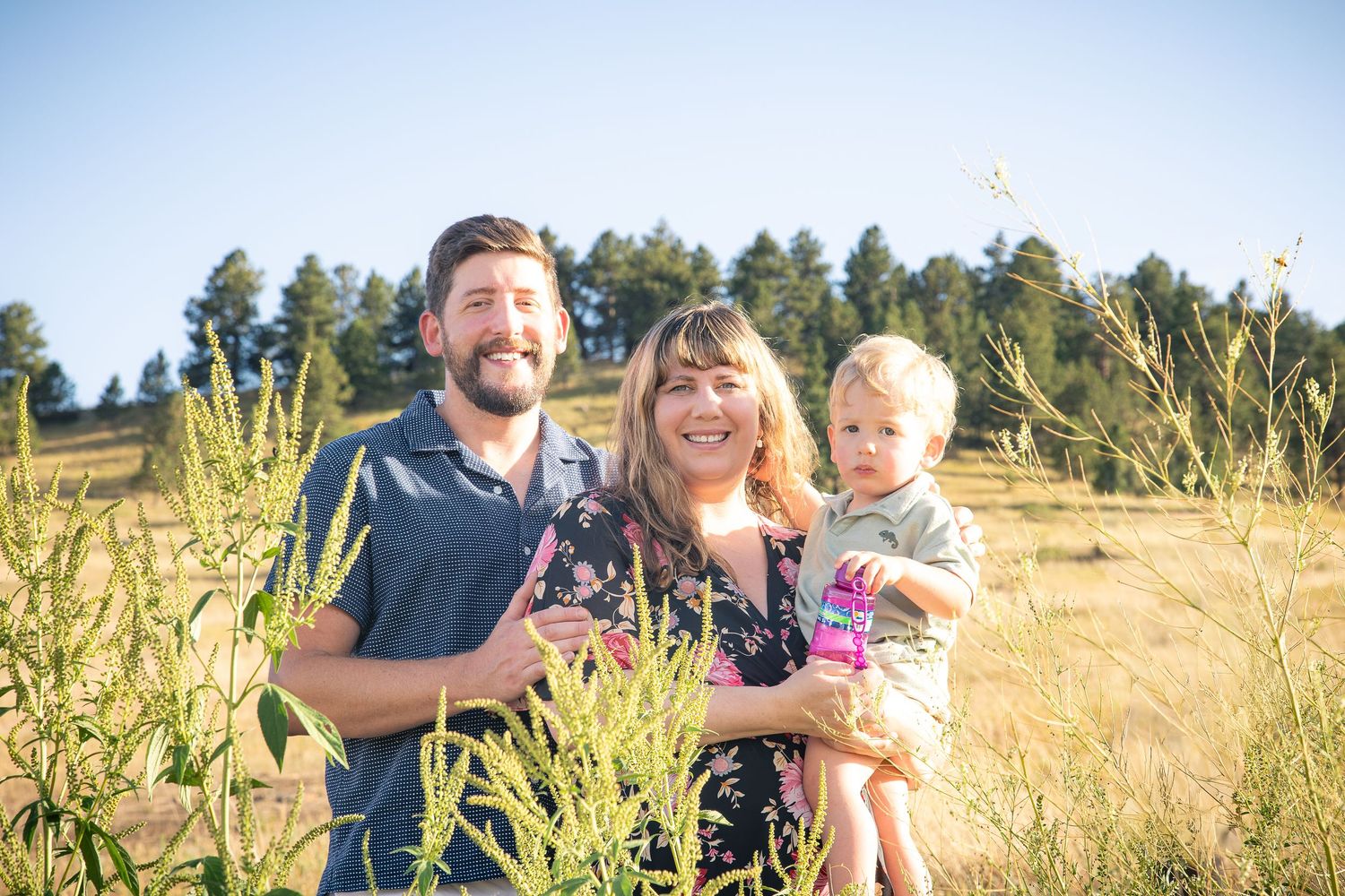 A happy family of three poses in a sunny meadow with pine trees, parents smiling with toddler in arms.