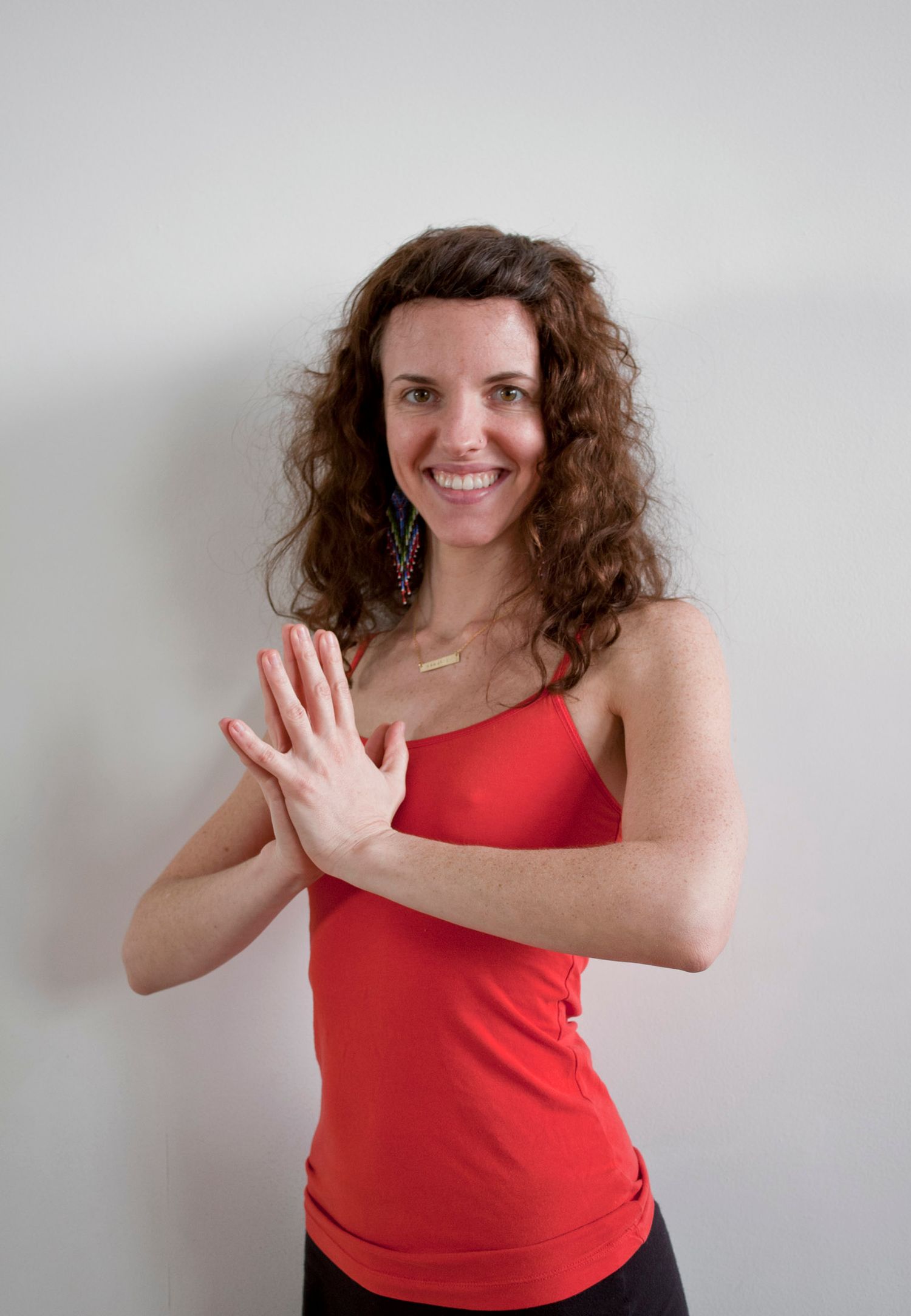 Smiling woman with curly hair in red tank top performing reverse prayer hand yoga pose.