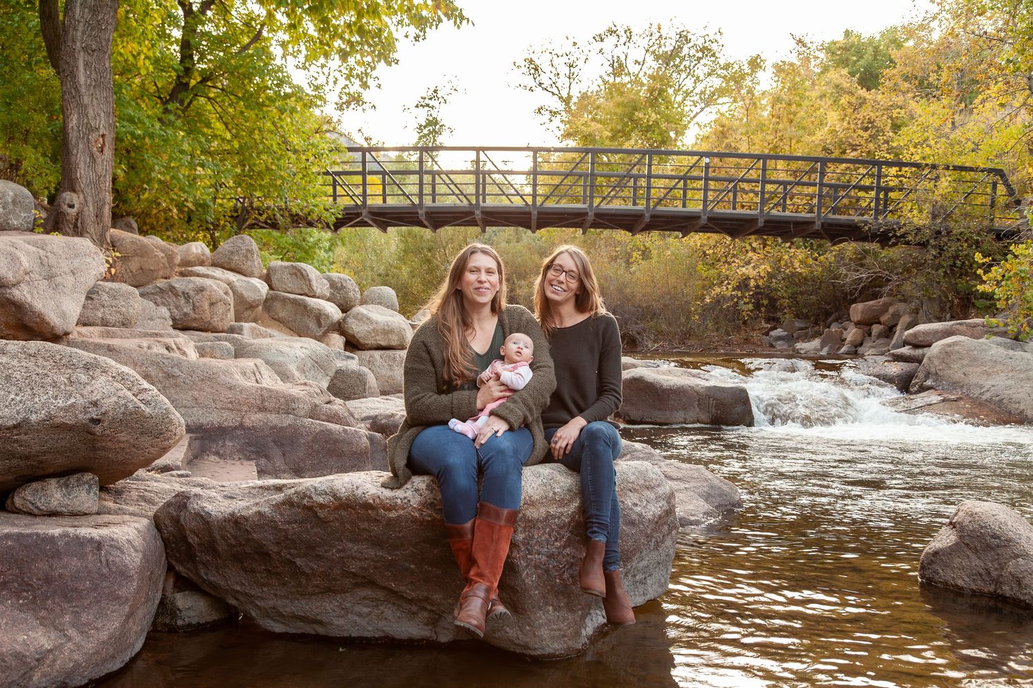 Two women sit on rocks by a flowing stream with a bridge and autumn trees in the background, one holding a baby.