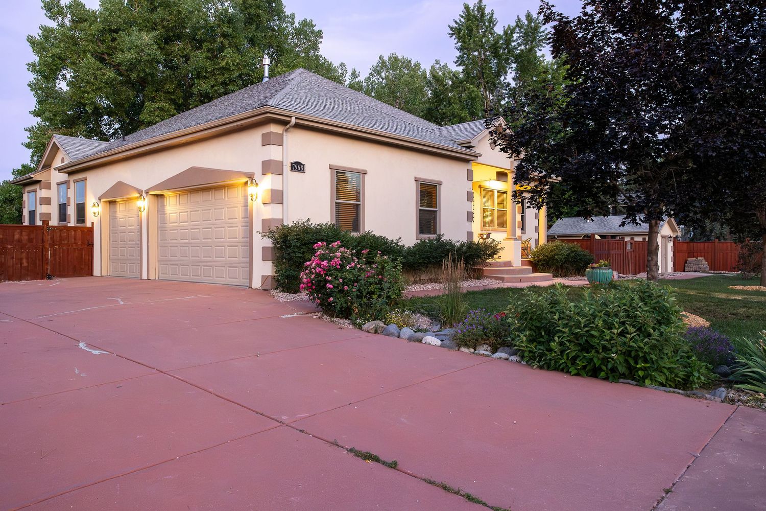 Charming beige stucco home with attached garage, lush trees, and blooming flowers in a well-maintained front yard at dusk.