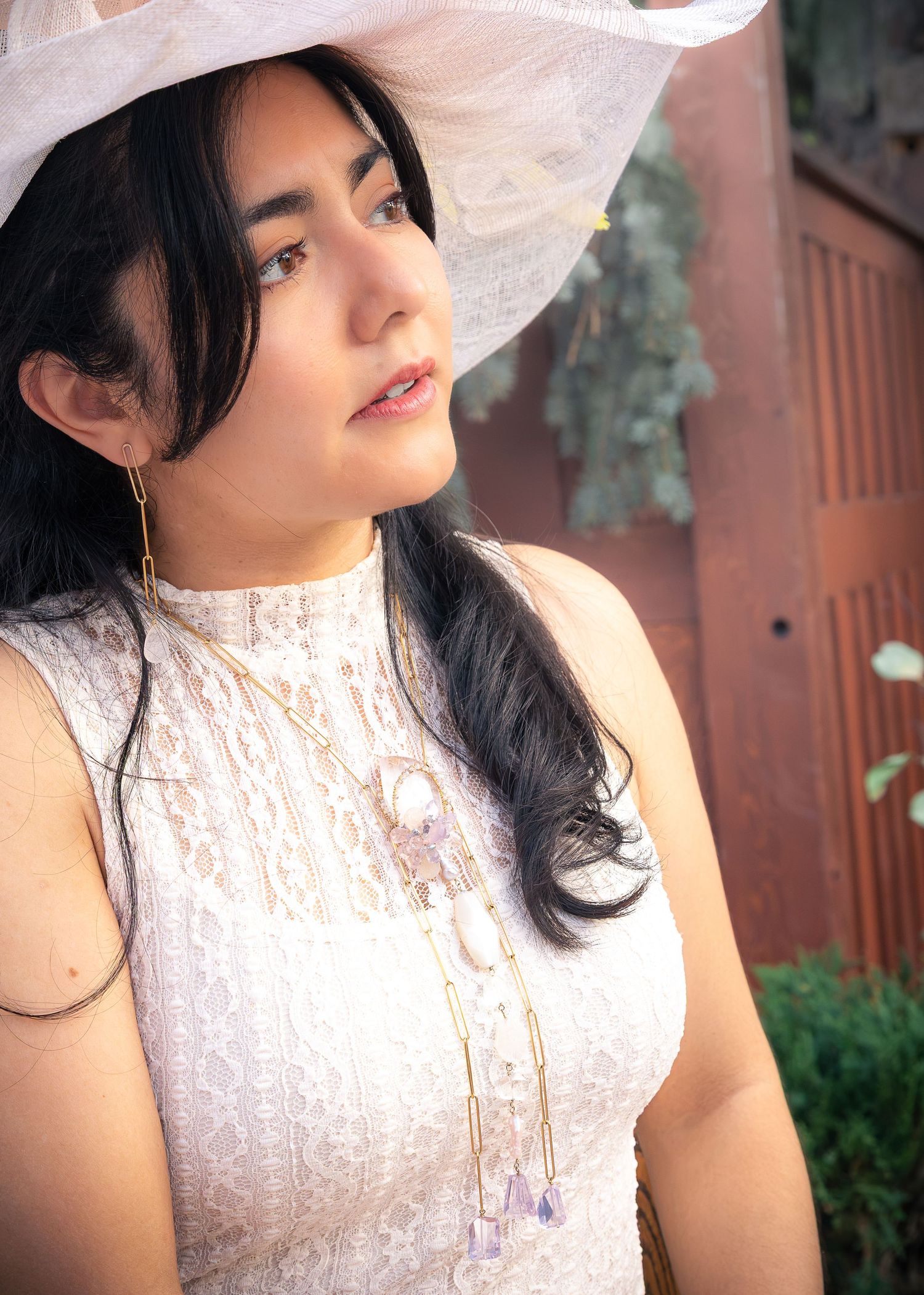 Woman in a white lace sleeveless top and wide-brim sun hat wears a long beaded pendant necklace, gazing aside near a rustic wooden gate.