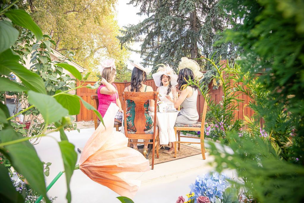 Four women in decorative hats sit and chat on wooden chairs in a sunny garden framed by green leaves and colorful flowers.