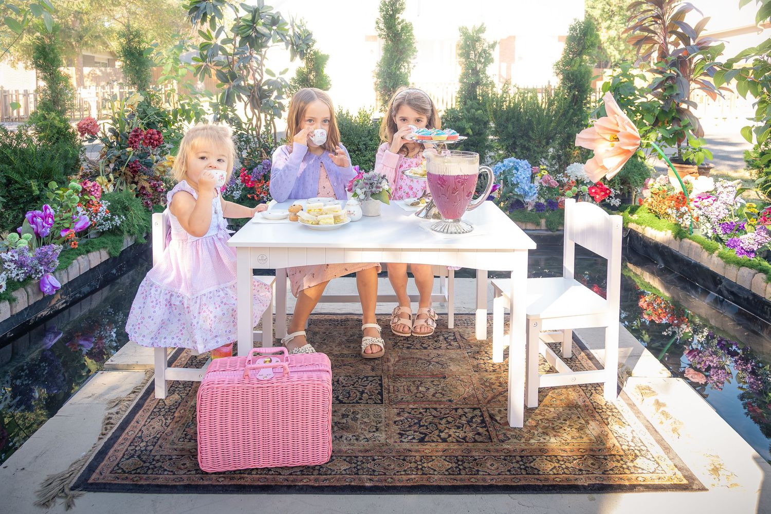Three young girls in pastel dresses enjoy a tea party at a white table in a flower garden, with a pink wicker basket on a patterned rug.