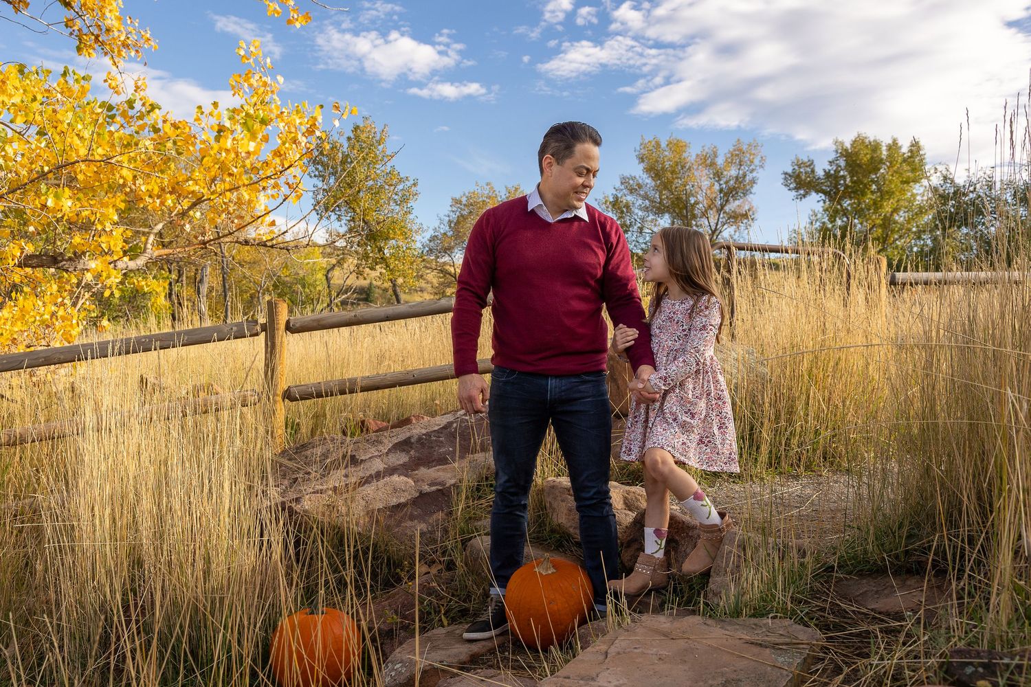 Father and daughter share a tender moment surrounded by pumpkins and fall foliage in a scenic outdoor setting.