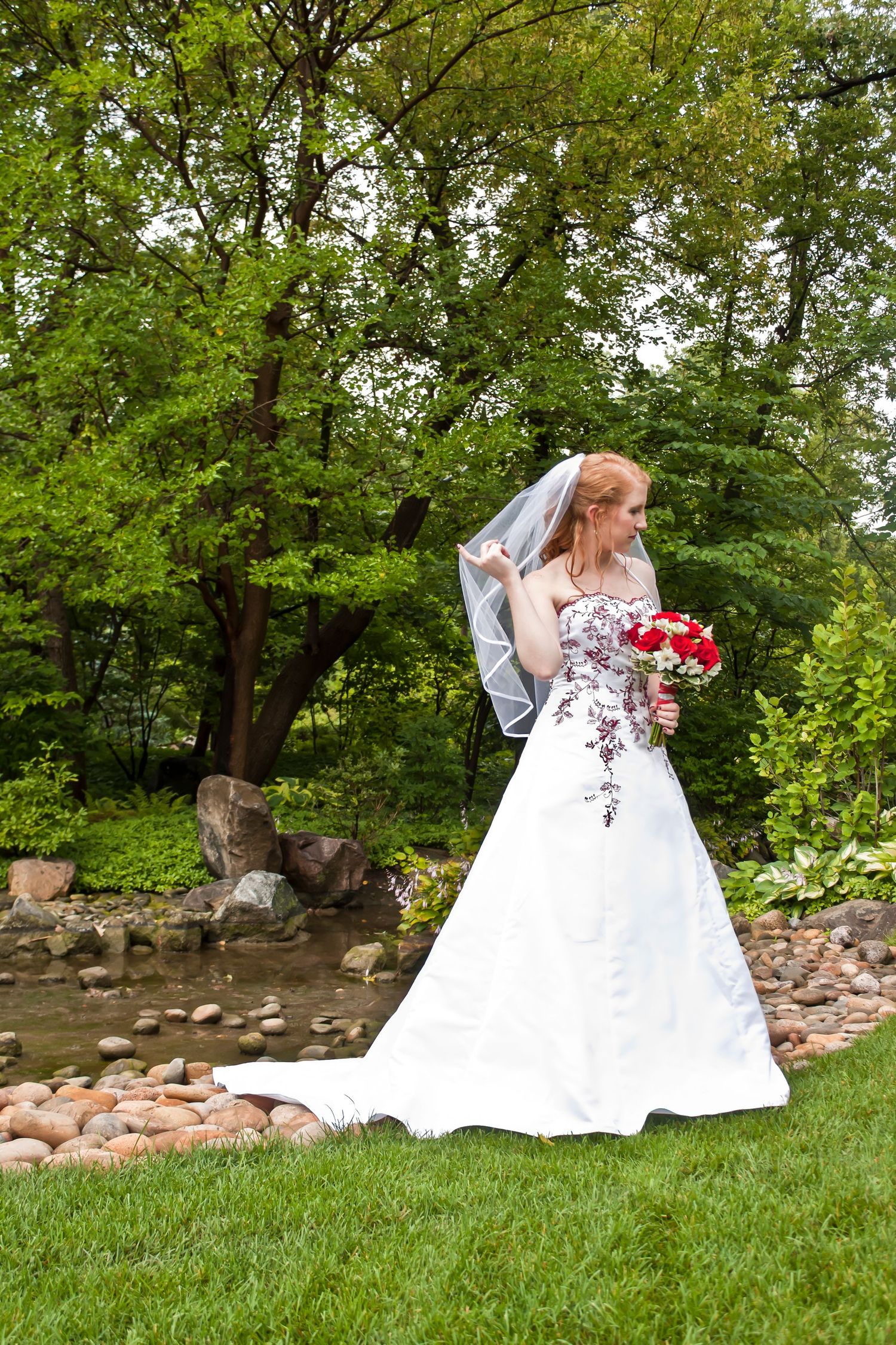 Red-haired bride in white gown holding red bouquet stands by a garden stream surrounded by lush green trees.