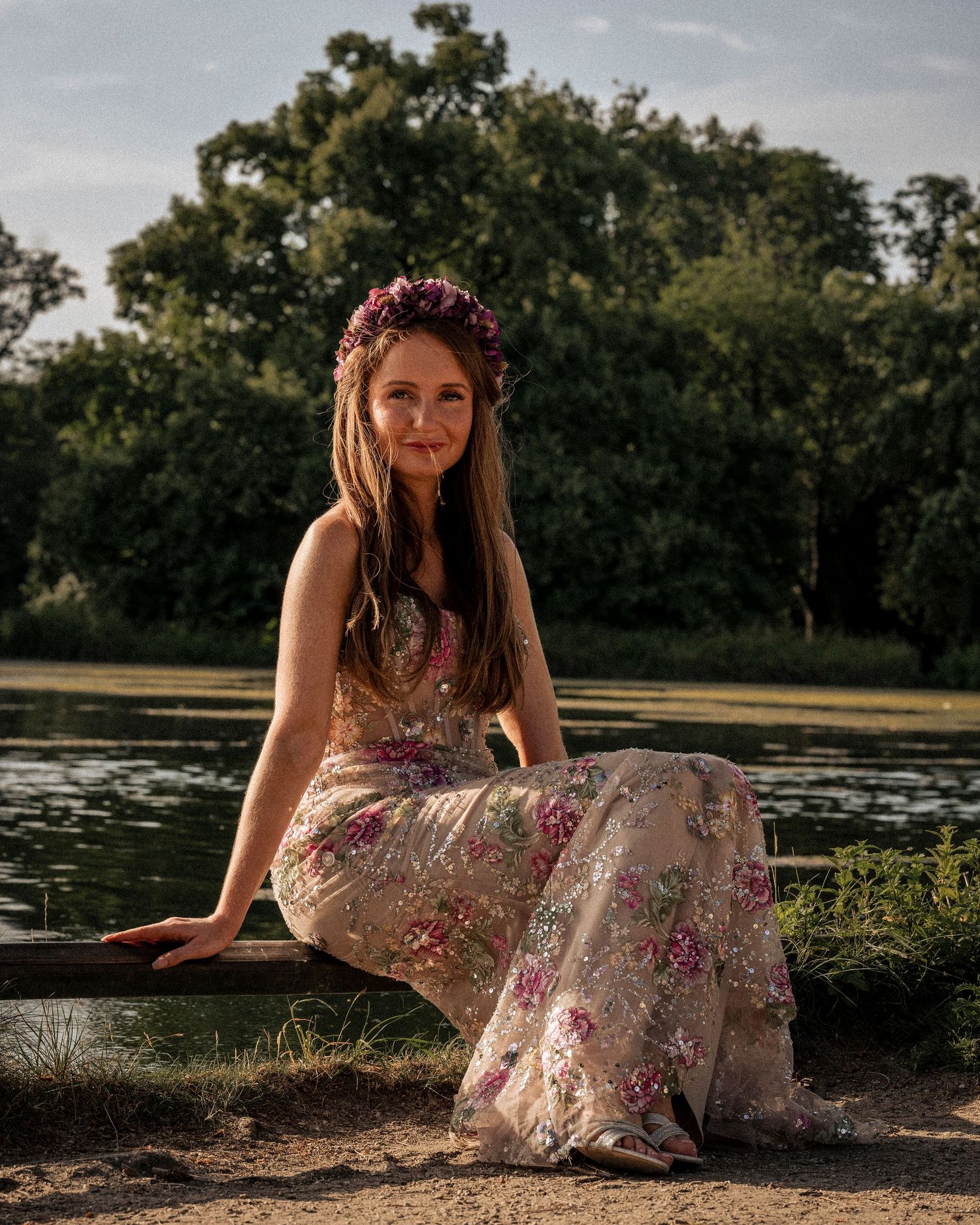 Young woman in floral dress and flower crown sits gracefully by a serene pond surrounded by lush green trees.