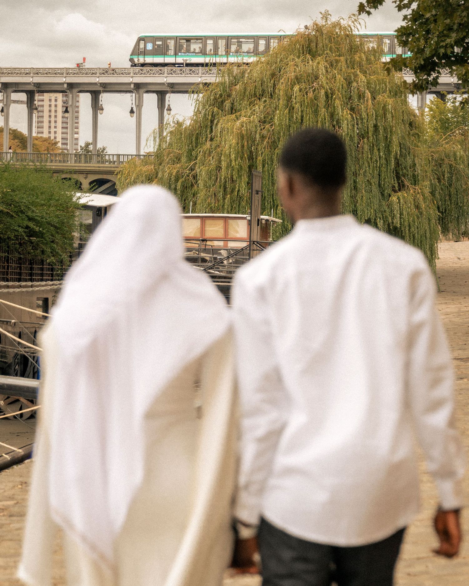 Couple in white attire walking near a willow tree with Paris metro bridge in the background on a cloudy day.