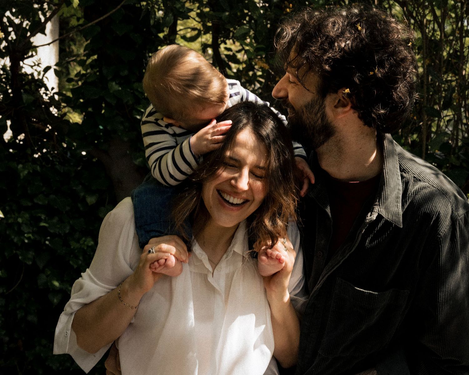 A smiling family with a toddler on mom's shoulders shares a warm embrace outdoors.