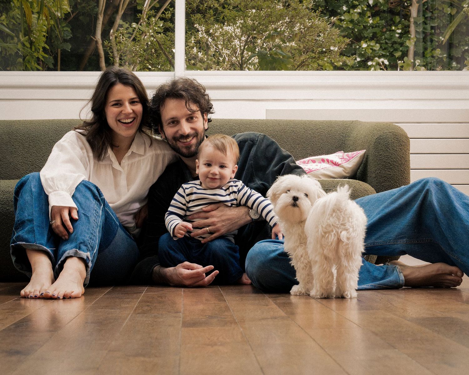 Happy family with baby and white fluffy dog sitting on floor by green sofa at home.