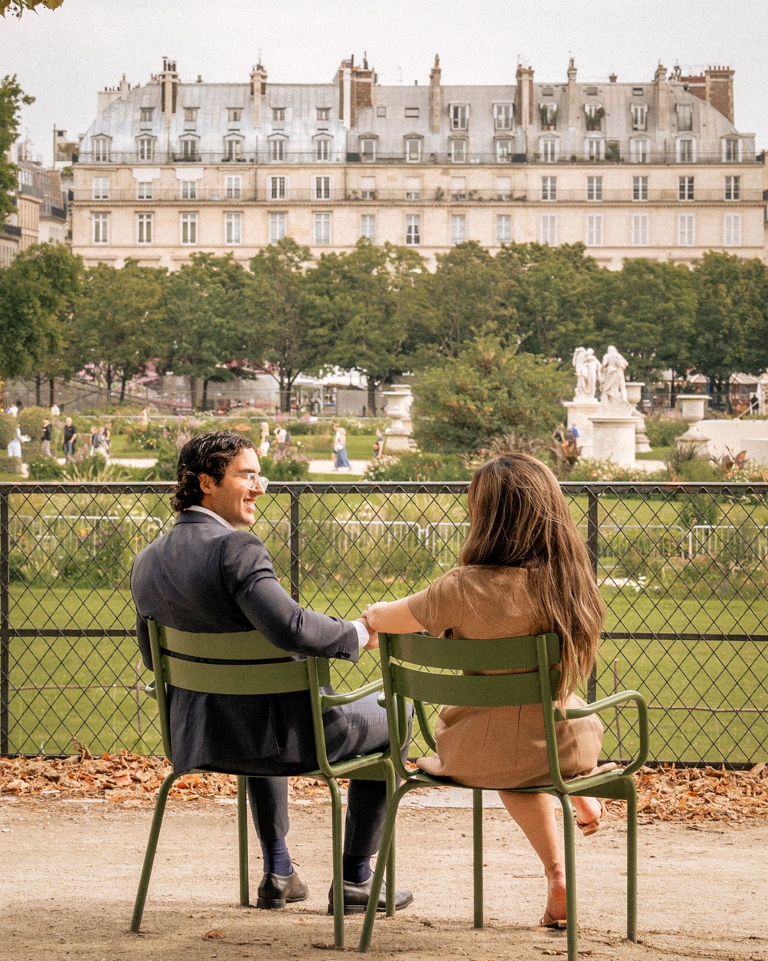 Couple sitting on green chairs in the Tuileries Garden, Paris, with Haussmann buildings in the background.