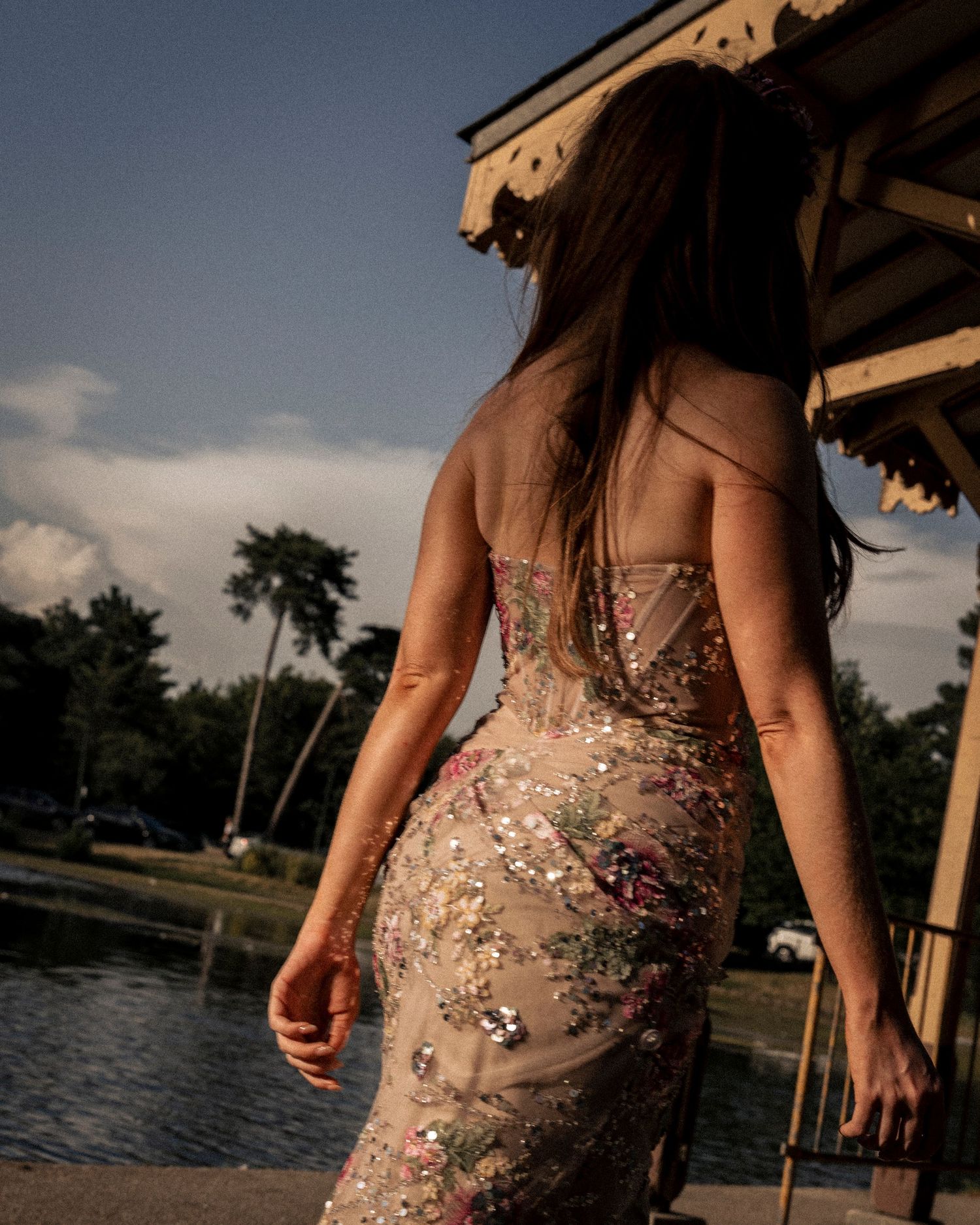 Woman in backless floral sequin gown stands near waterfront gazebo at dusk.
