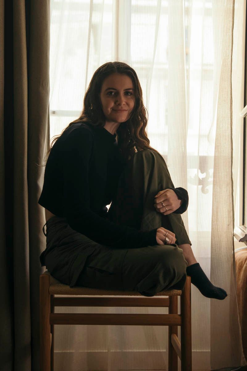 Young woman sitting cross-legged on wooden chair by sheer curtained window indoors.