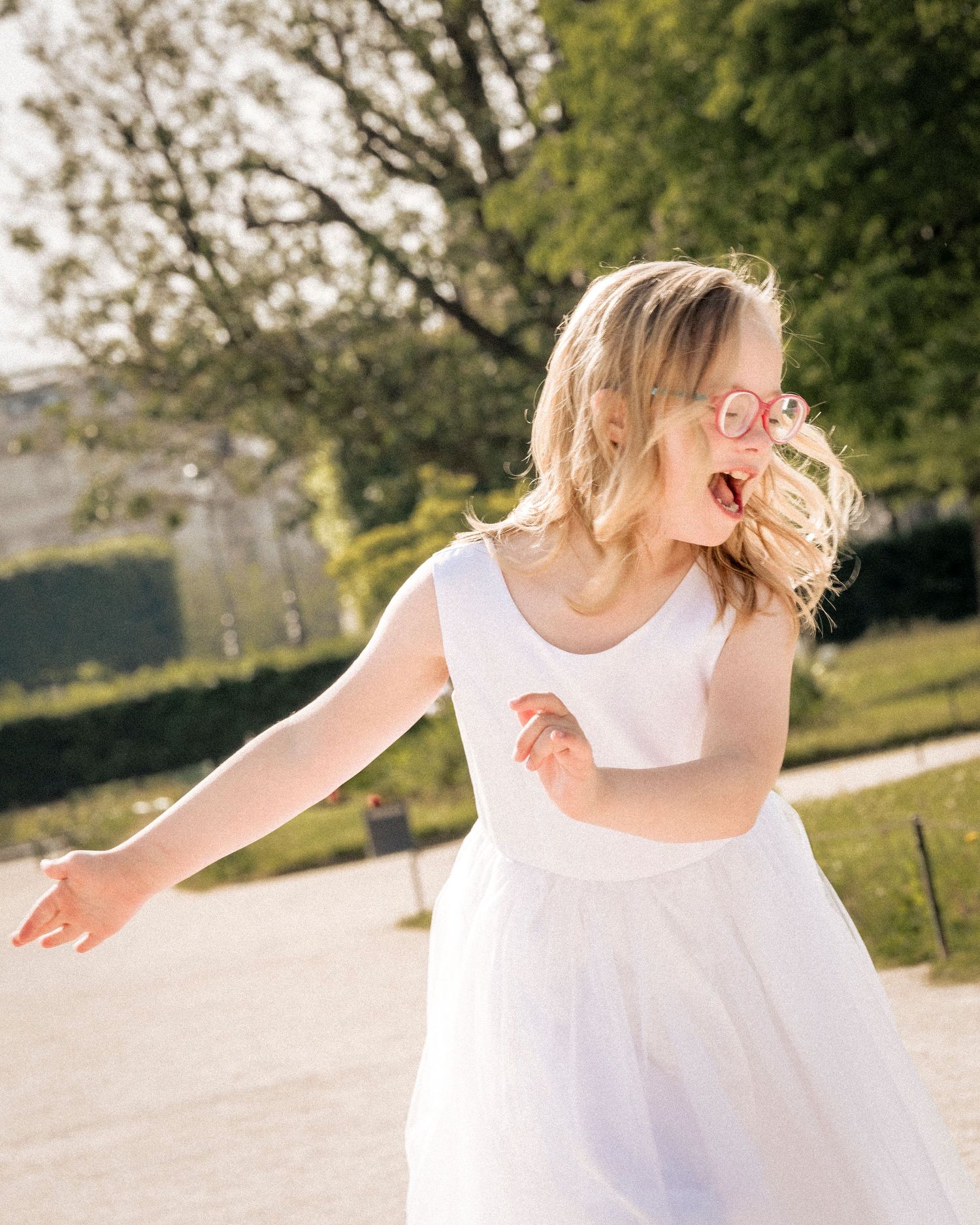 A young girl in a white tulle dress and pink sunglasses dances playfully in a park.