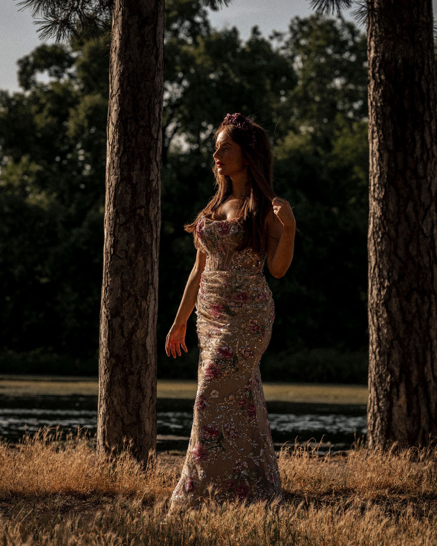 Young woman in floral gown poses dramatically between pine trees at golden hour.