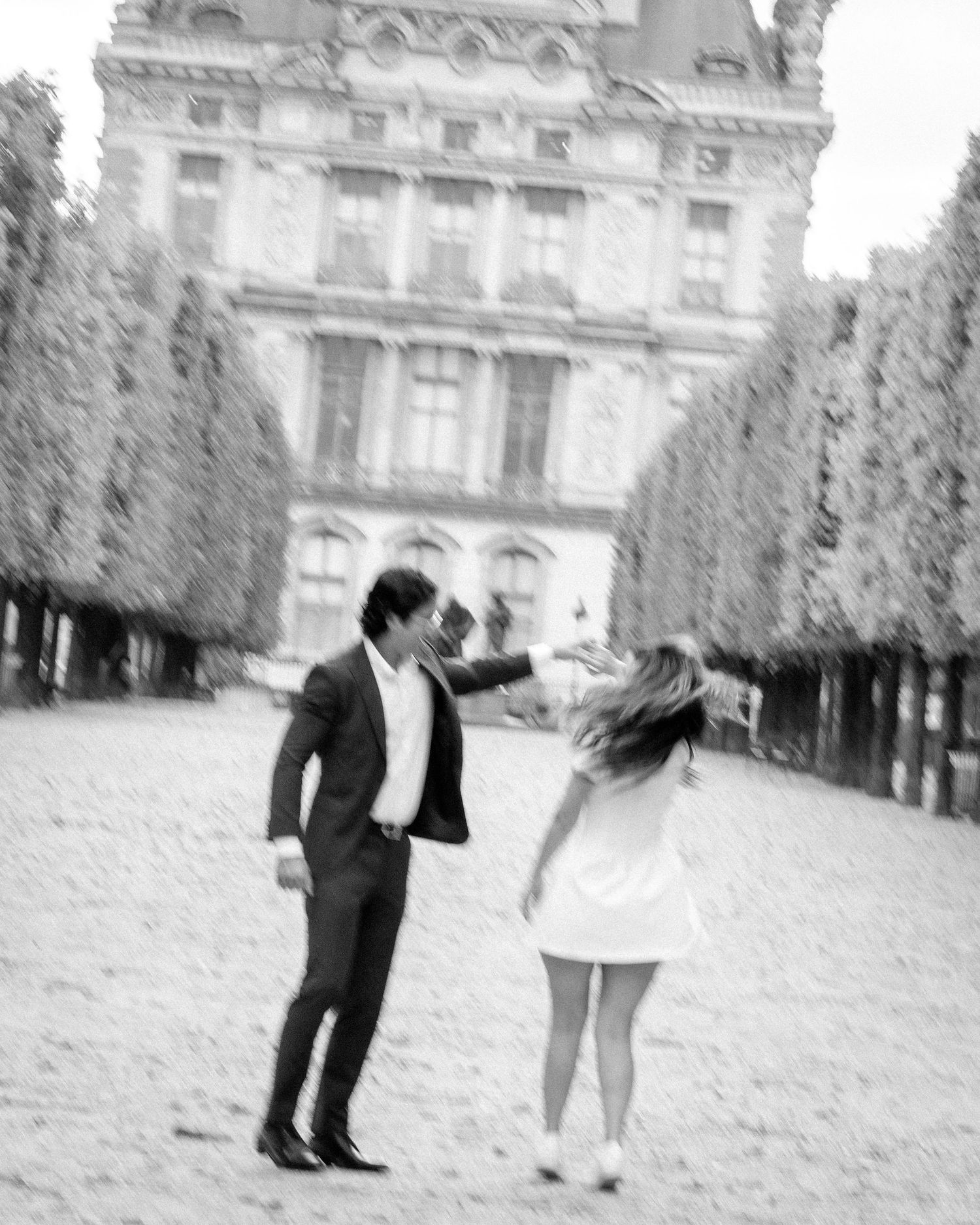 Blurry black and white photo of a couple dancing on a gravel path in Paris with a grand building behind them.