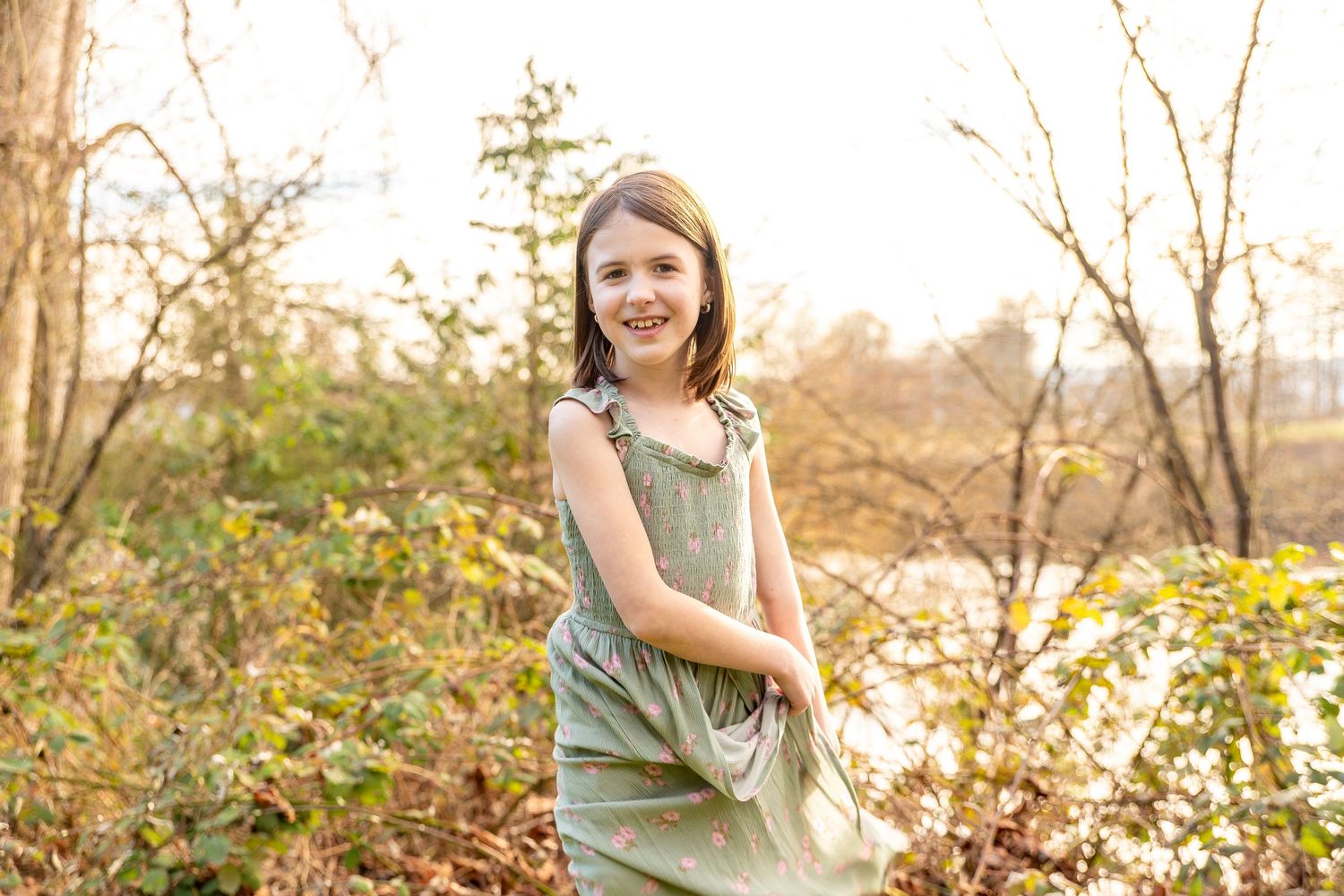 A person in an olive green dress poses outdoors among fall foliage during golden hour lighting.