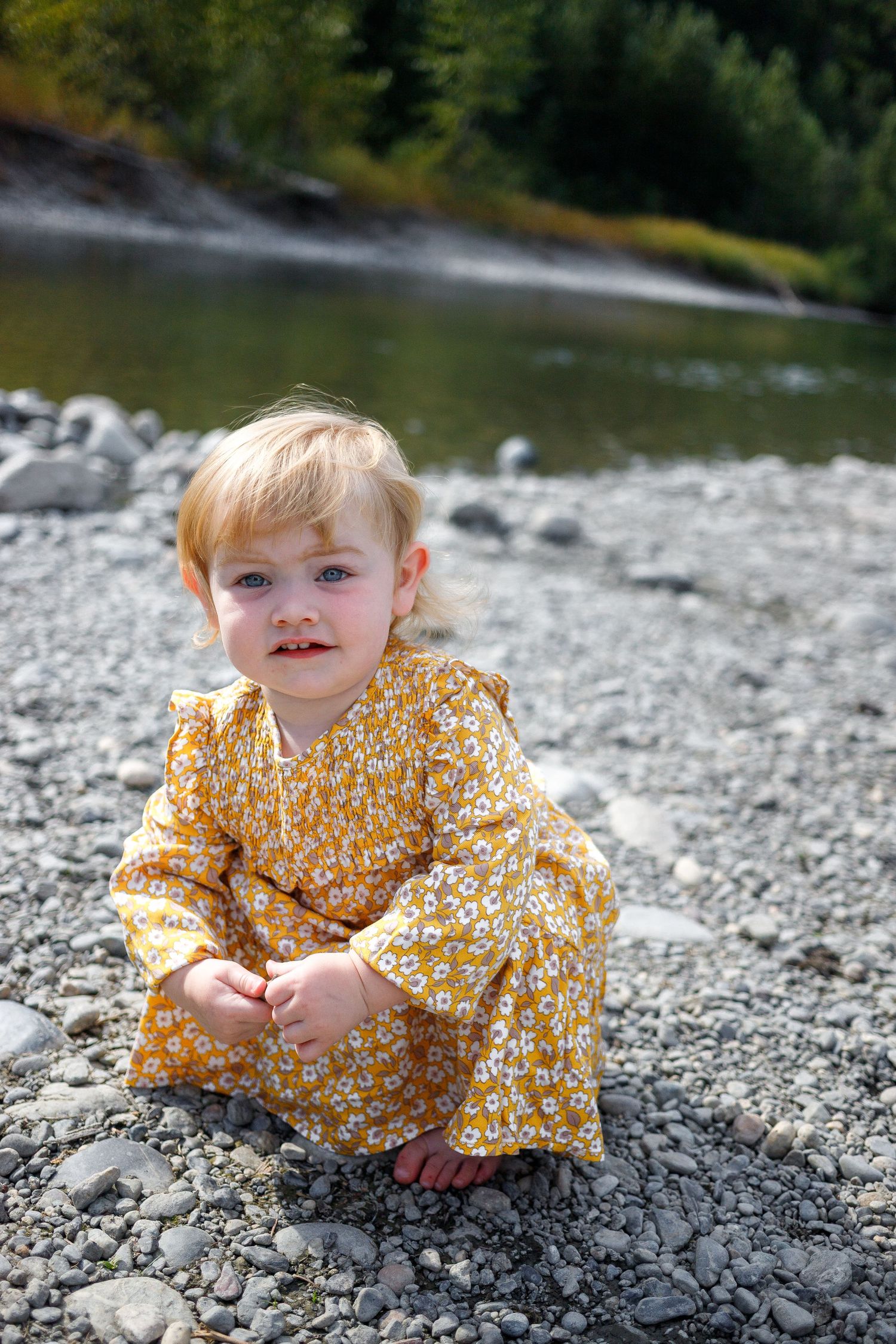 A small child wearing a yellow floral dress sits and plays on a rocky riverbank on a sunny day.