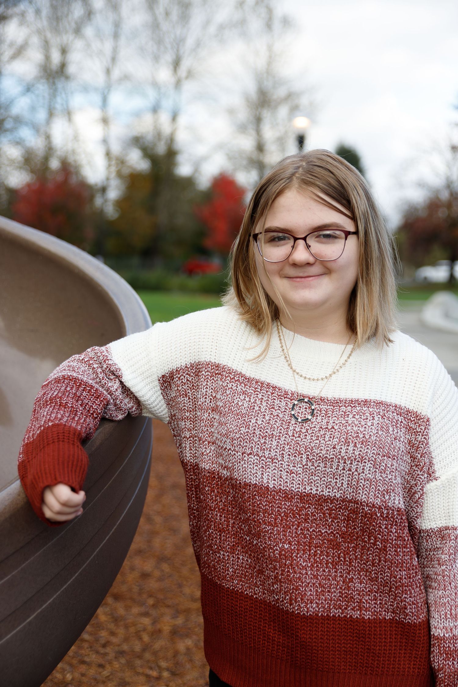 Someone in a color block sweater with glasses poses near a playground slide on an autumn day.