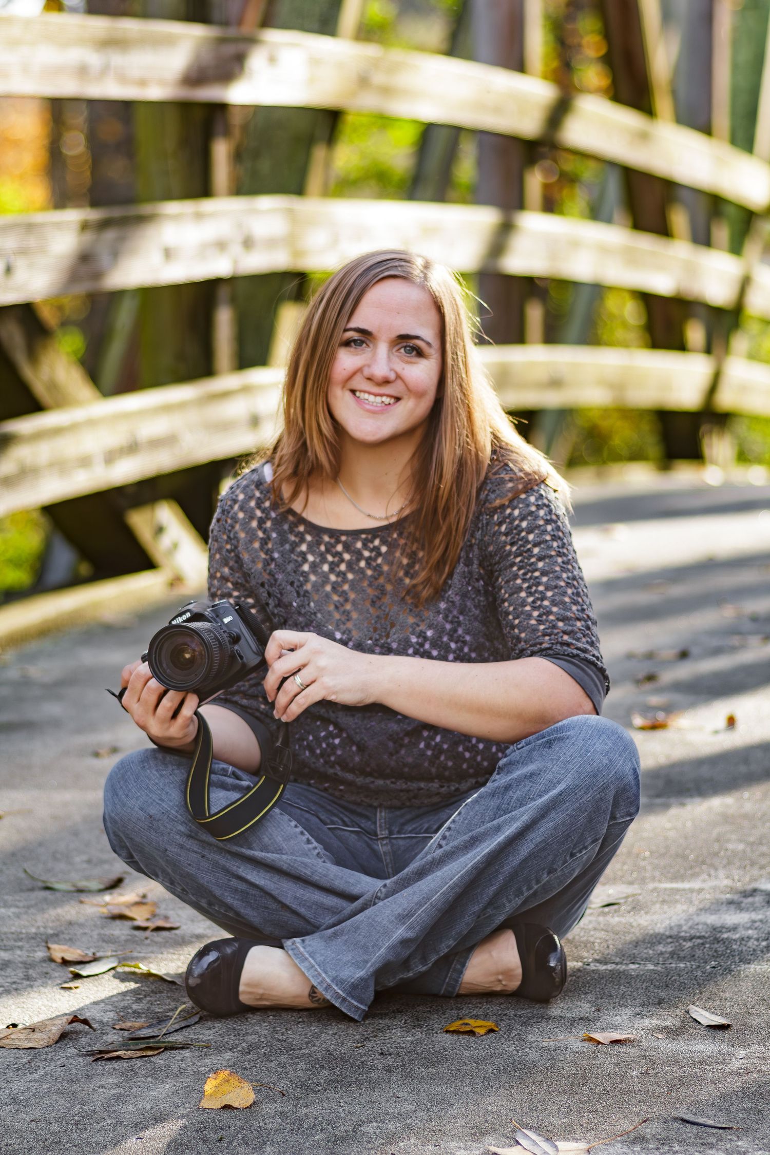 A photographer sits cross-legged on wooden bridge while holding DSLR camera outdoors in casual clothing.