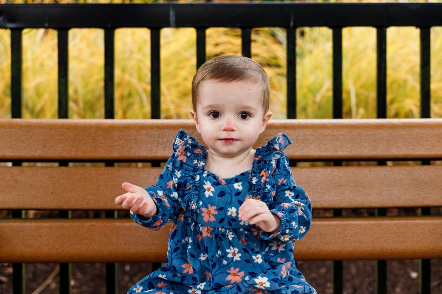Small child wearing a blue floral dress sits on a wooden bench in front of a yellow fence.