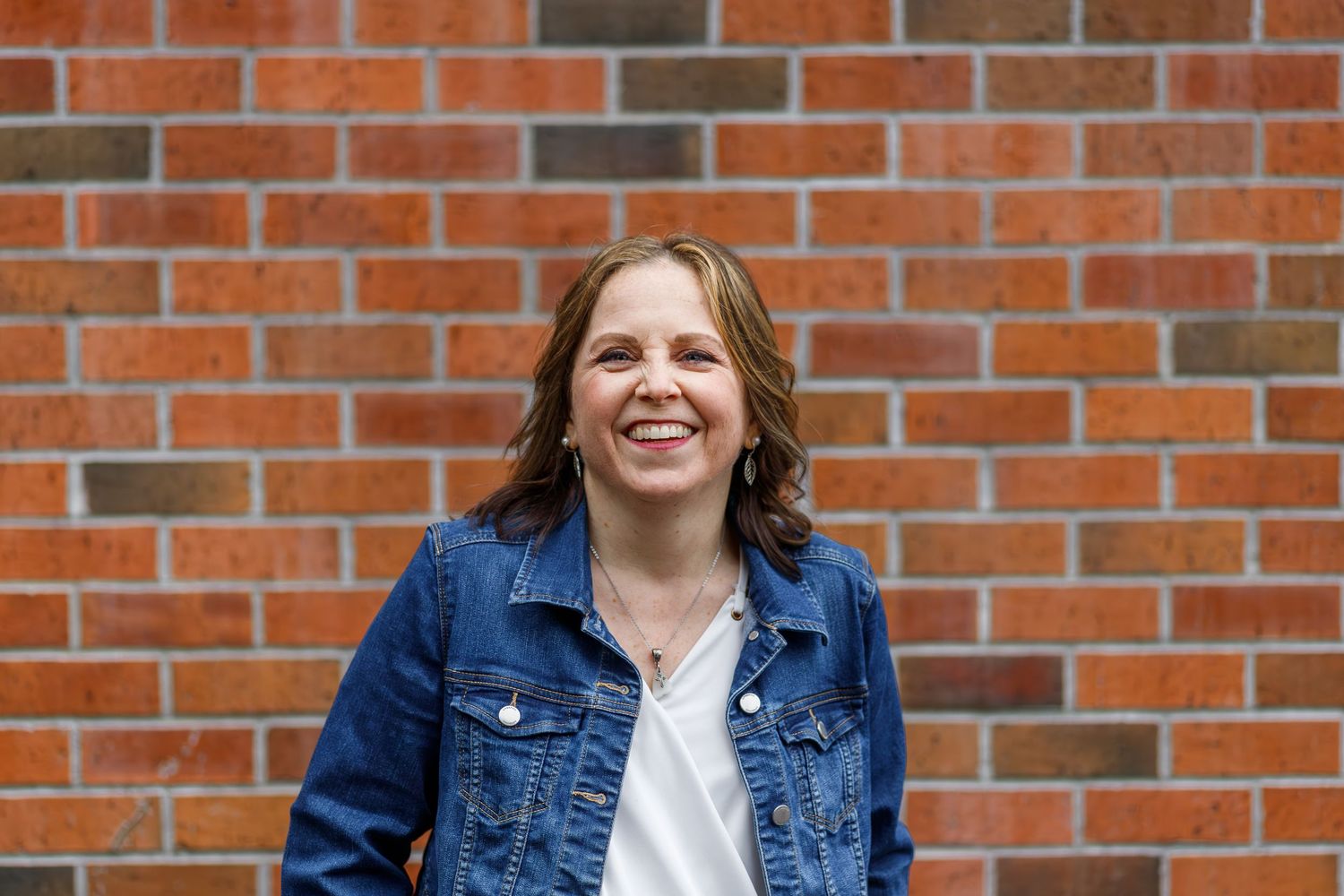 A smiling person wearing a blue denim jacket stands in front of a red brick wall.