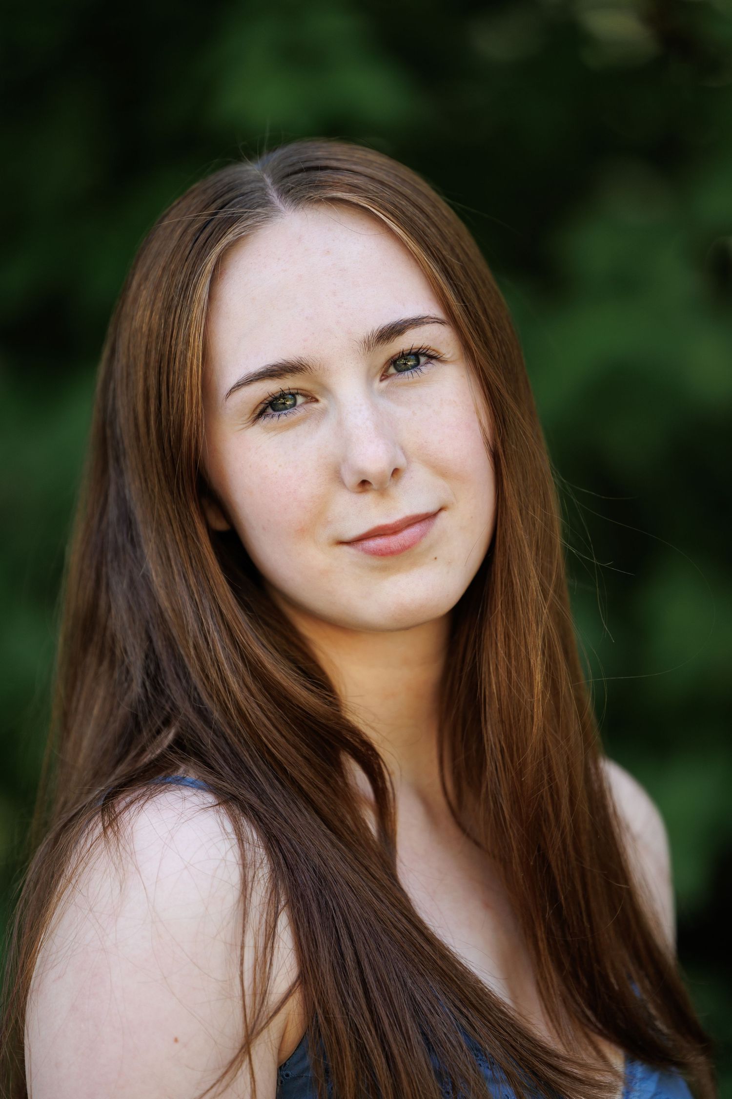 Young woman with long auburn hair and green eyes smiling outdoors with green foliage background.