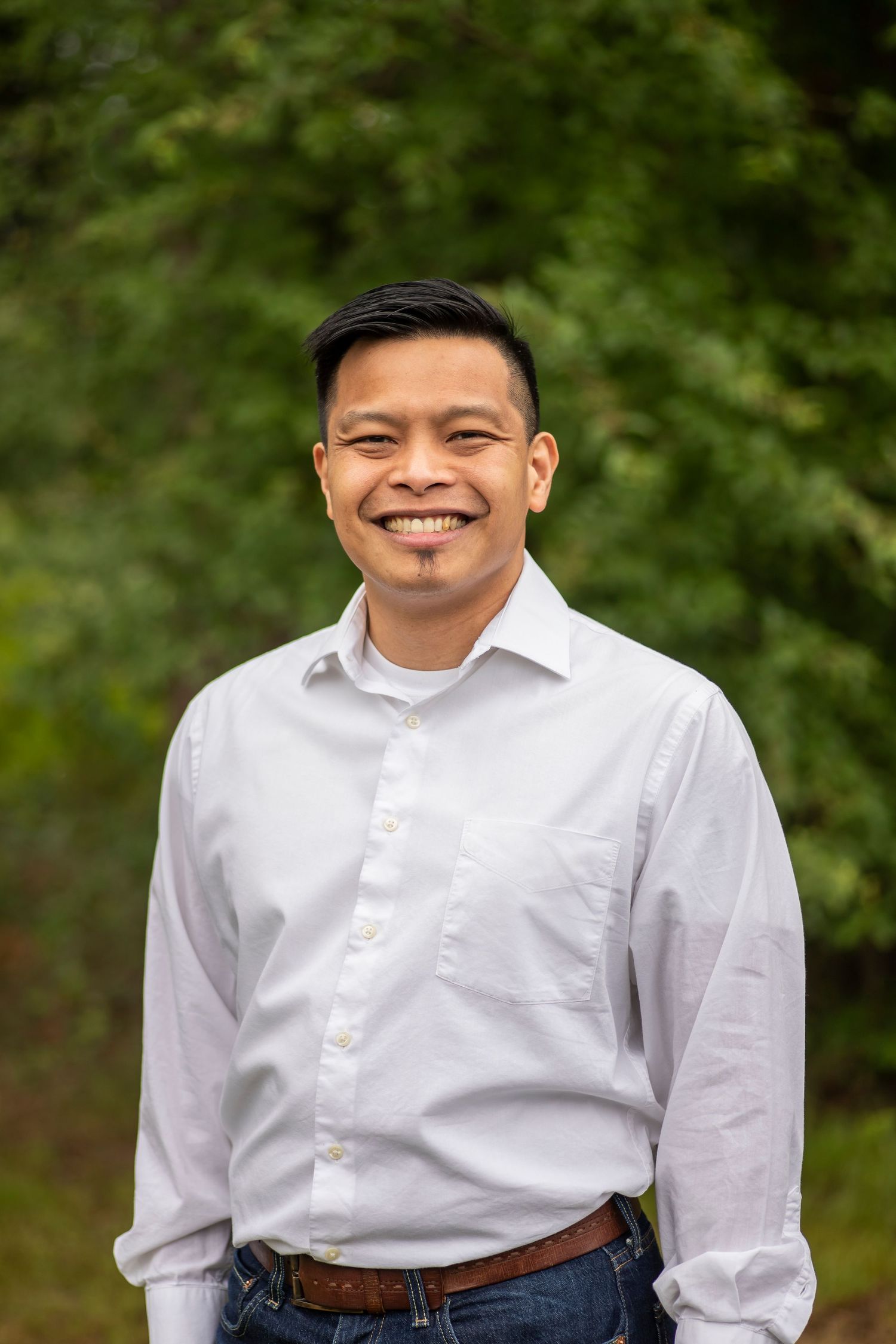 Person wearing a crisp white button-down shirt and brown leather belt smiles warmly against a blurred green outdoor background.