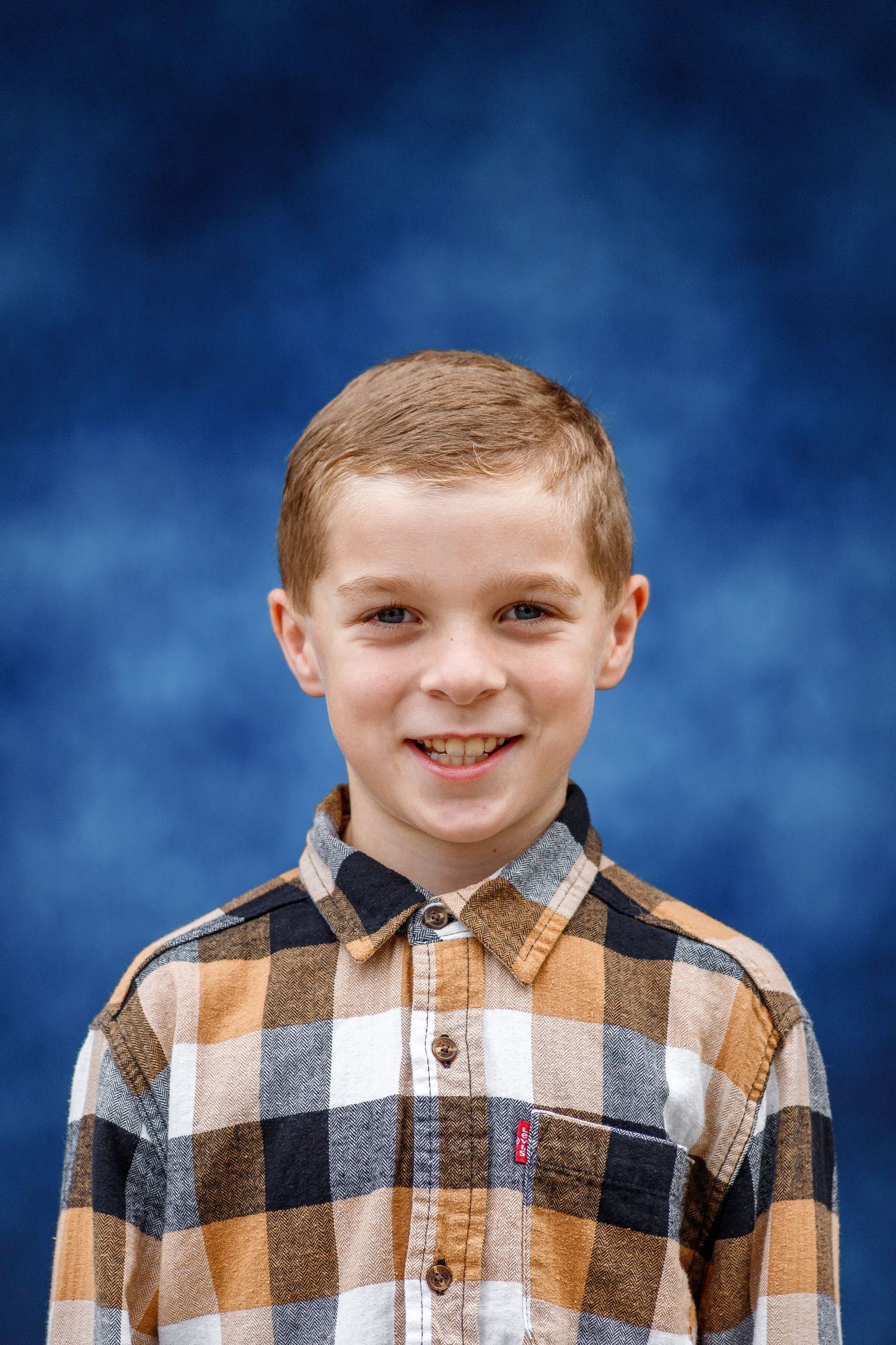 A smiling child wearing a brown and white buffalo plaid button-up shirt against a blue background.