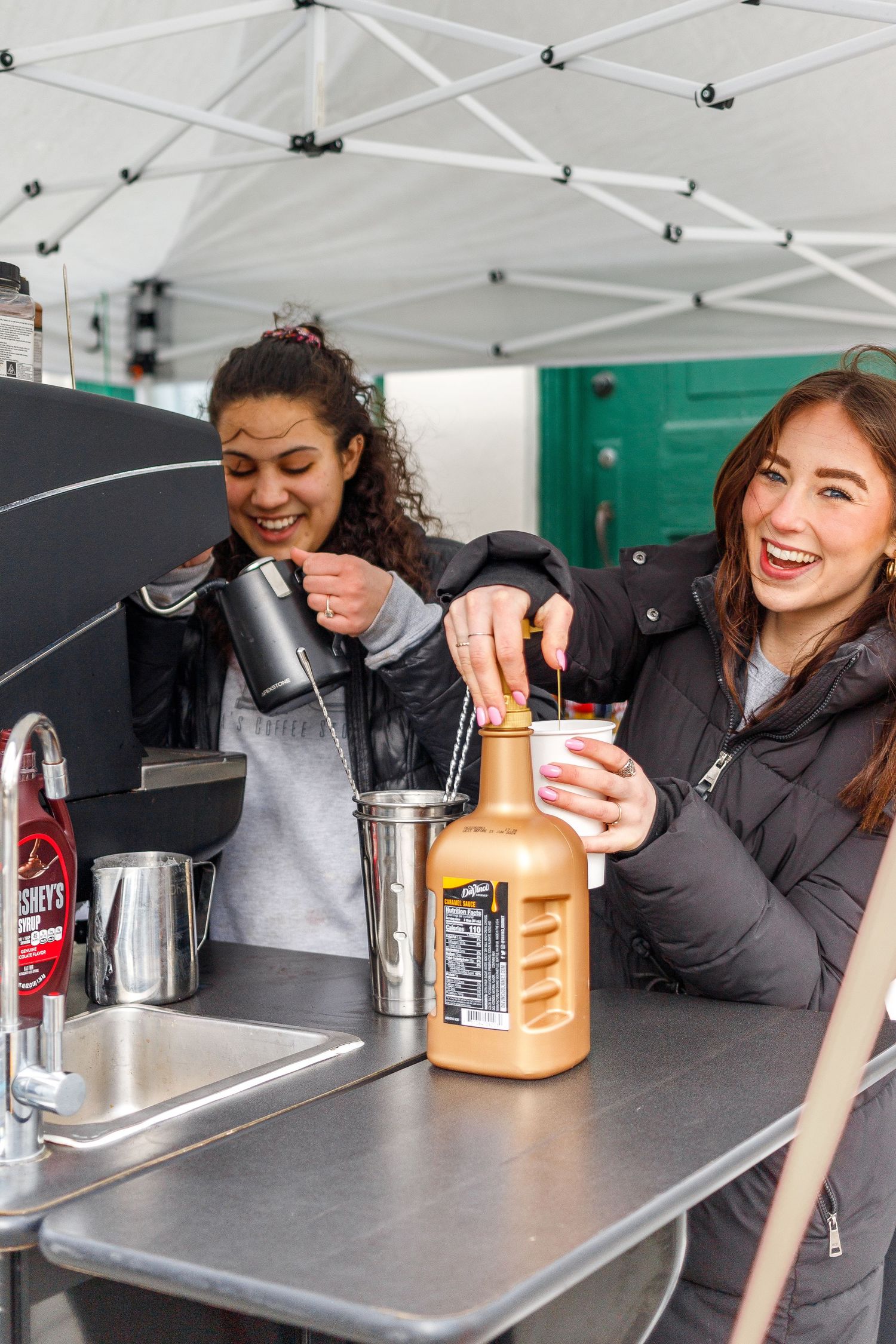 Baristas prepare beverages at an outdoor coffee stand with equipment and bottles on a stainless steel counter.