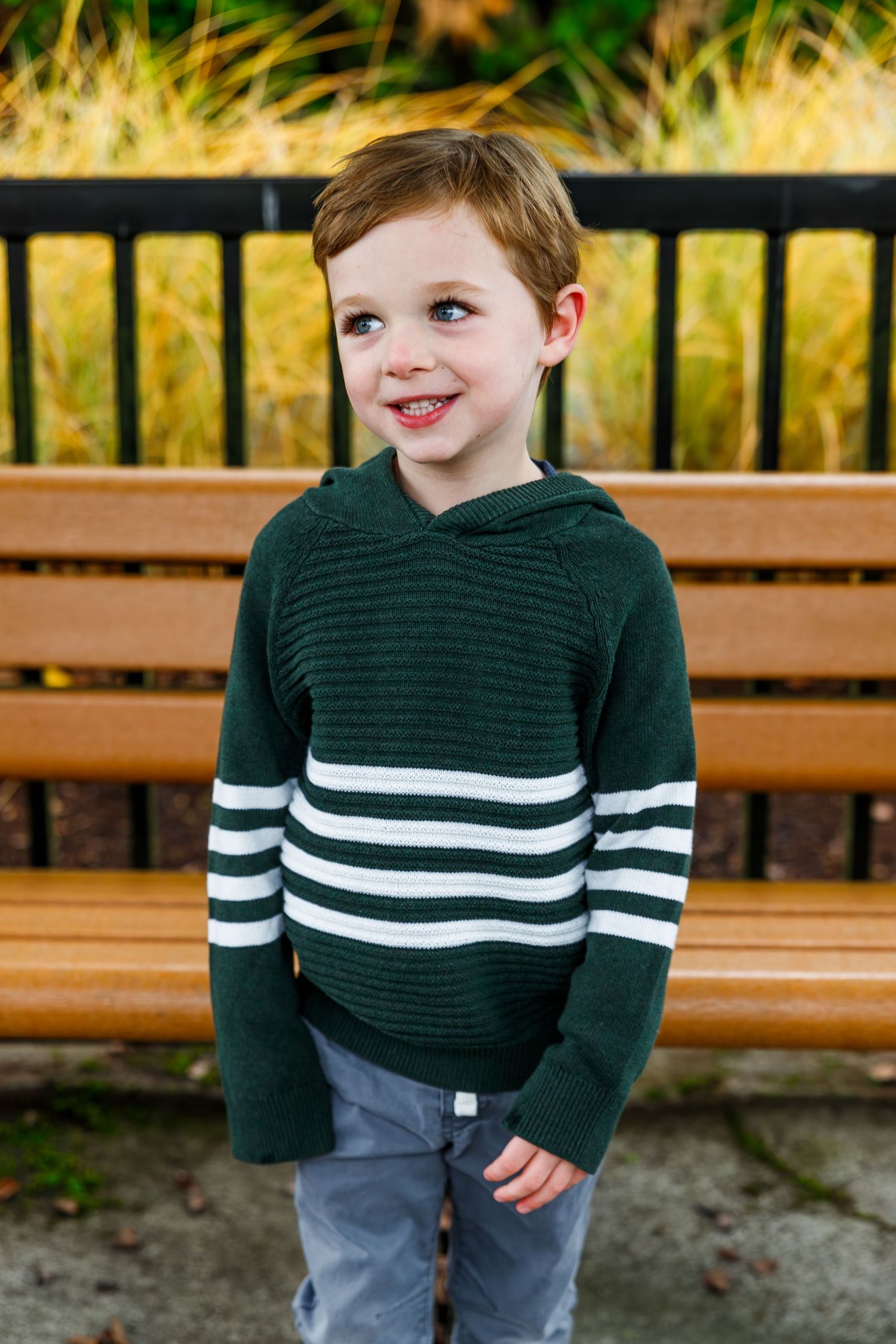 Young child smiles while wearing a green and white striped sweater with grey pants on wooden bench outdoors.