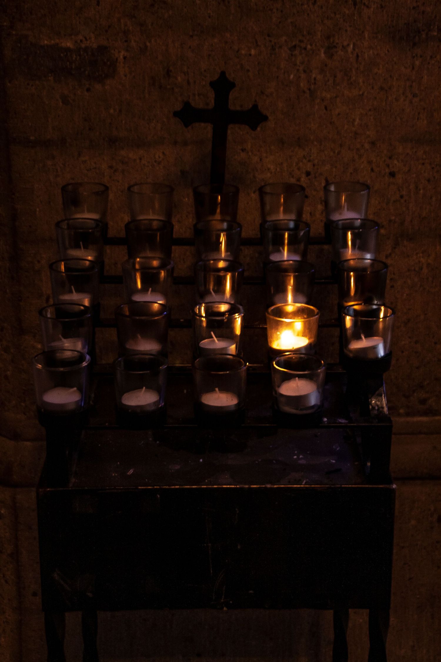 Prayer candles arranged in rows with a cross on a dark wooden stand in a church setting.