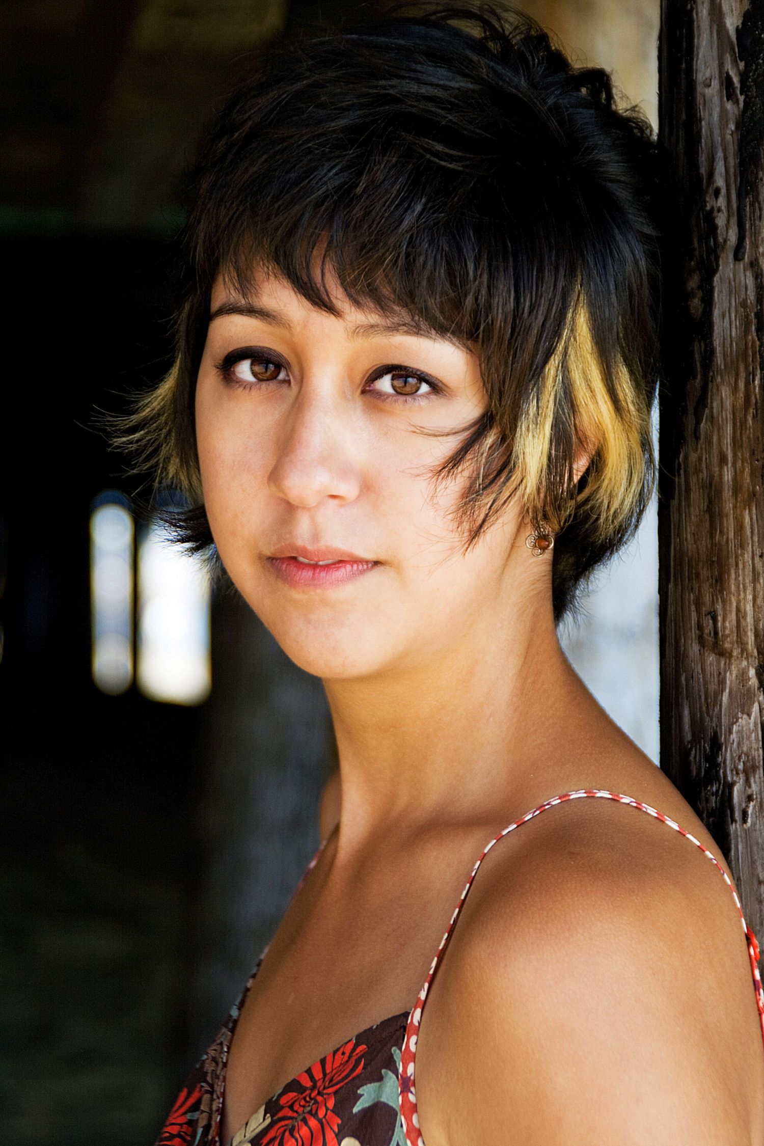 Portrait of a person with short black and blonde hair wearing a red patterned top against a dark background.